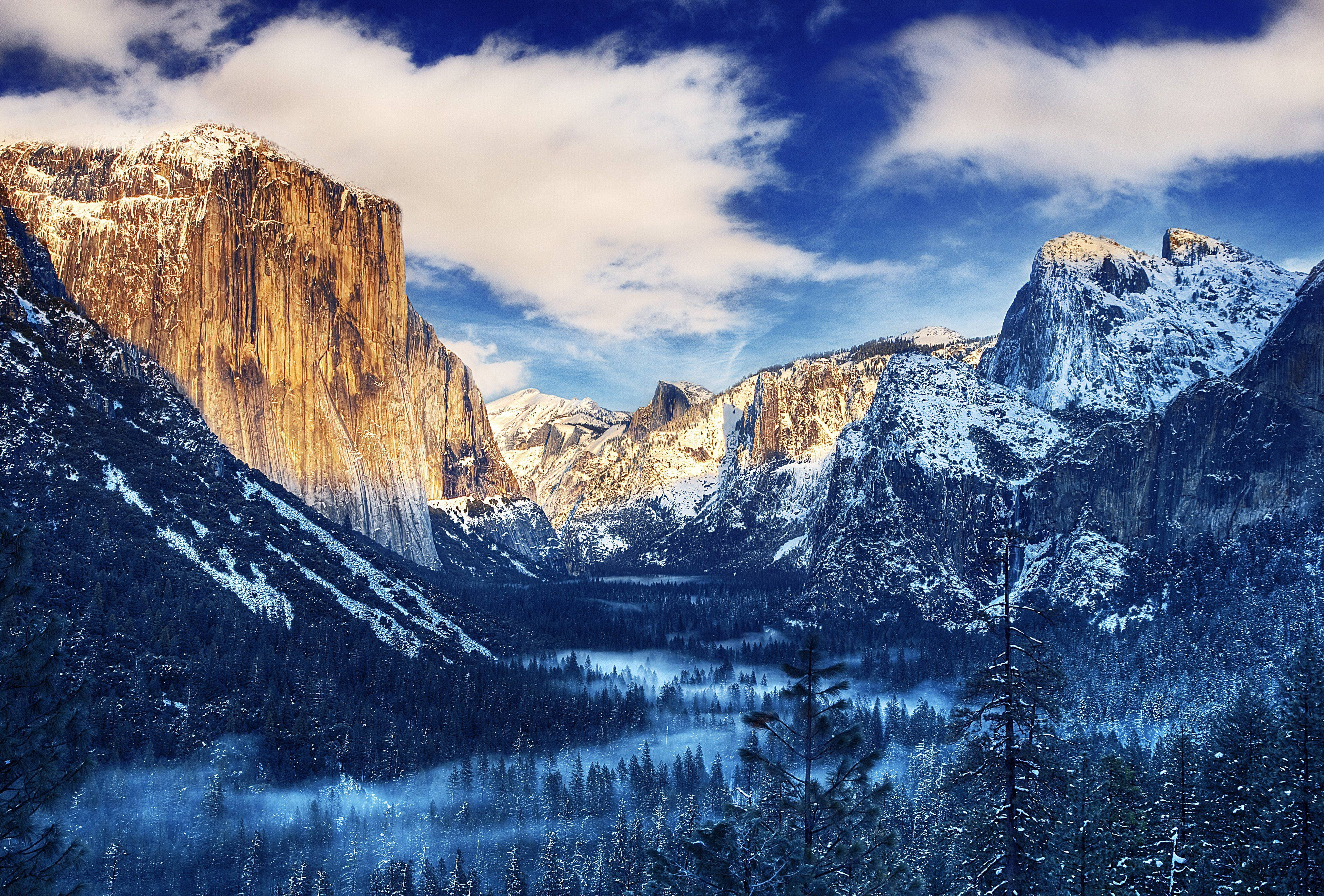 macOS 桌布 壁紙 — 白雪覆蓋的花崗岩懸崖聳立在一個充滿常綠樹木的霧氣山谷之上，天空湛藍，雲朵蓬鬆。