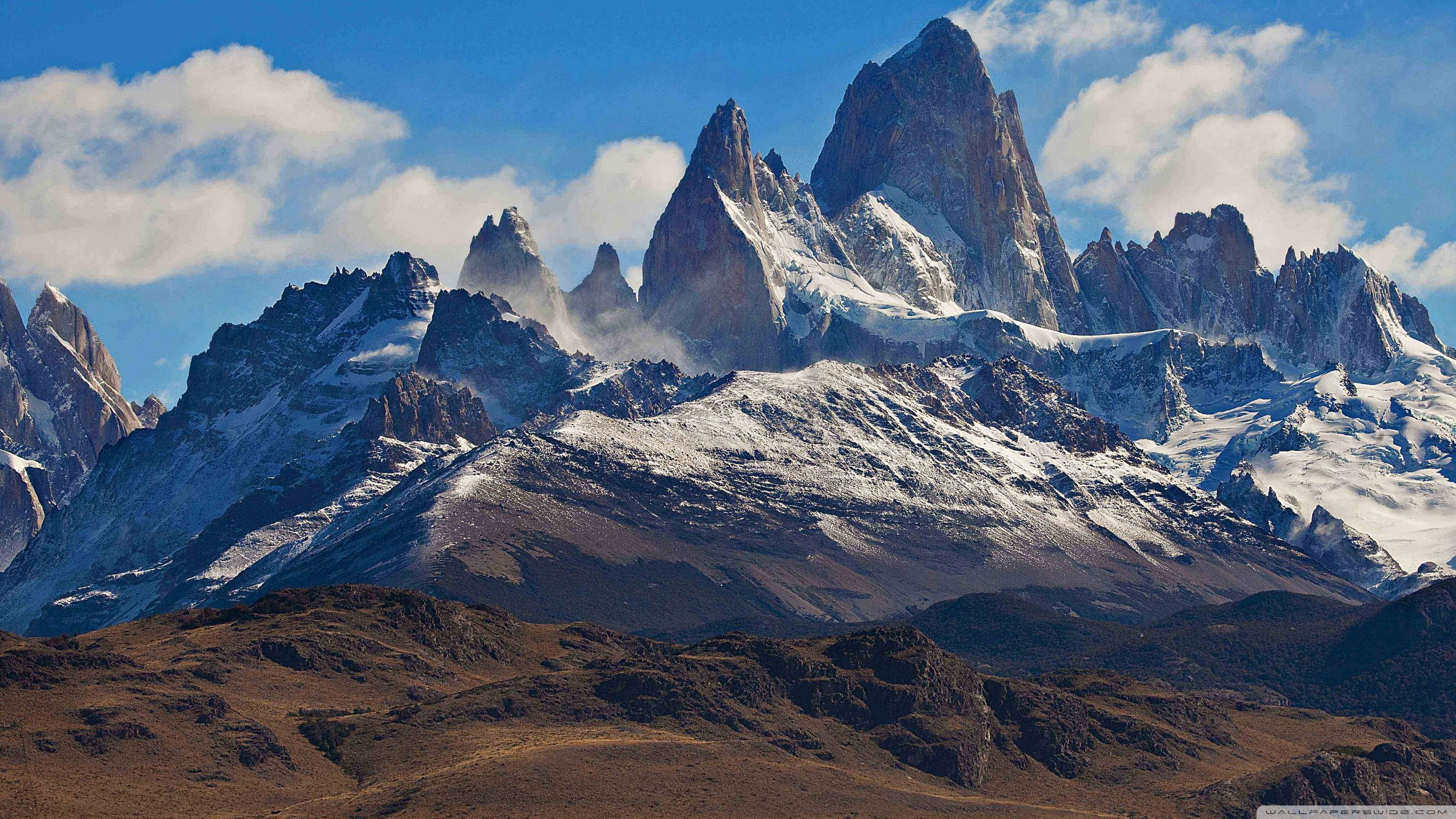 Patagonia 桌布 壁紙 — 白雪覆蓋的鋸齒狀山峰刺穿明亮的藍天，天空中有蓬鬆的白雲，下方是乾燥的棕色山麓。