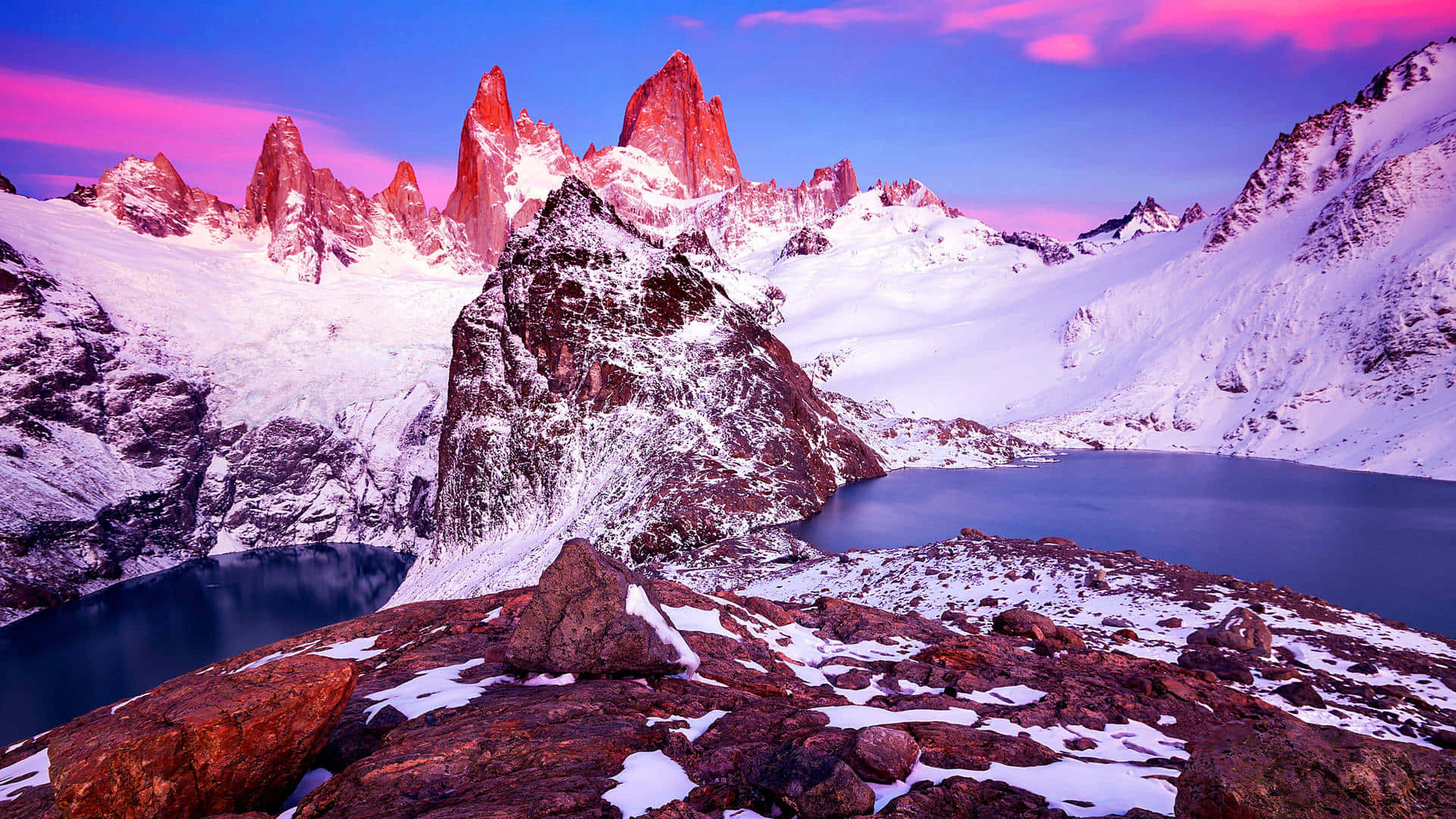 Patagonia 桌布 壁紙 — 白雪覆蓋的鋸齒狀山峰在日落時分散發出粉紅色和紫色的光芒，映照在由岩石地形環繞的寧靜高山湖泊之上。