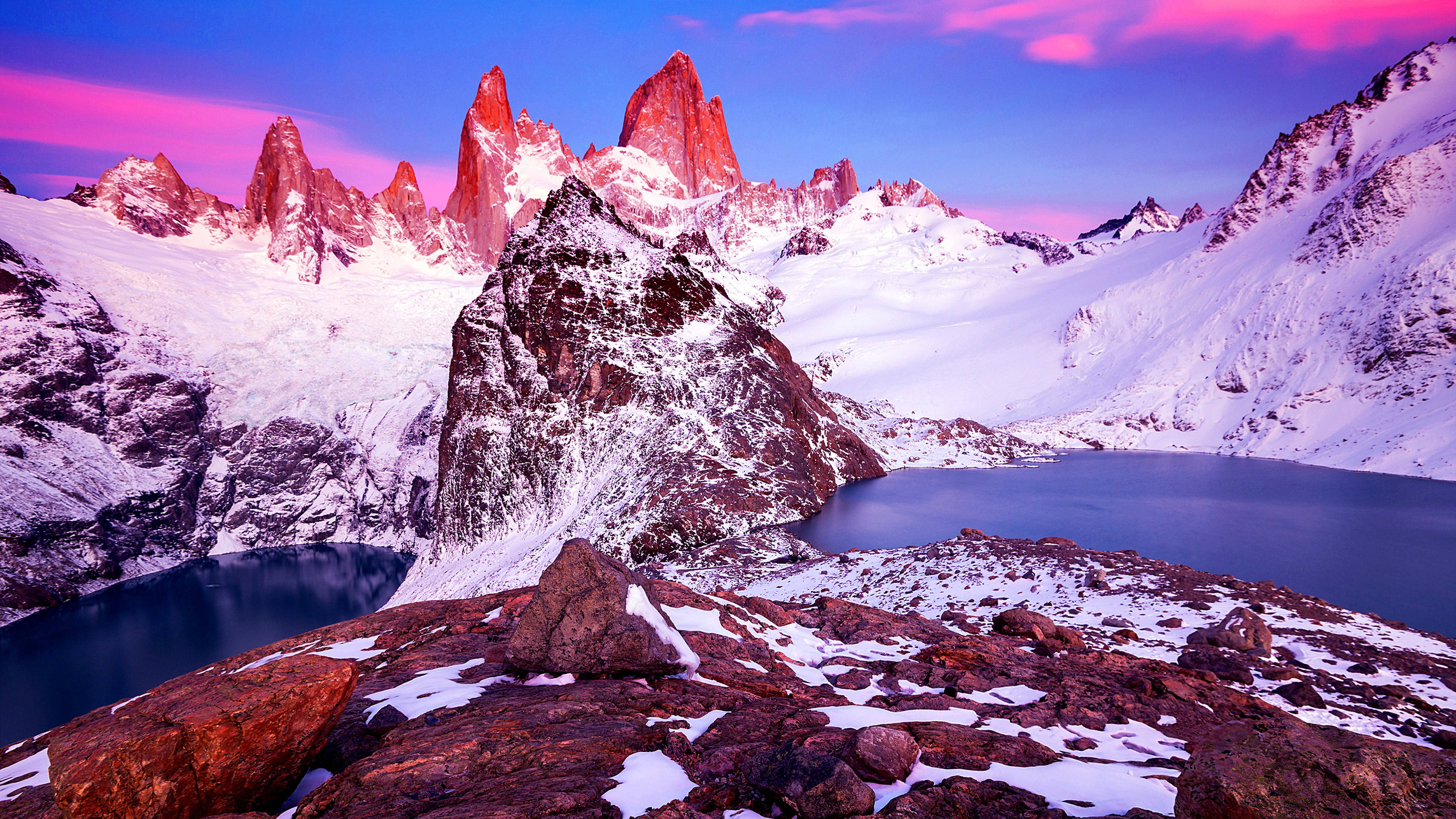 Patagonia 桌布 壁紙 — 白雪覆蓋的鋸齒狀山峰在日落時分散發出粉紅色和紫色的光芒，下方是平靜的高山湖泊，周圍是岩石地形。