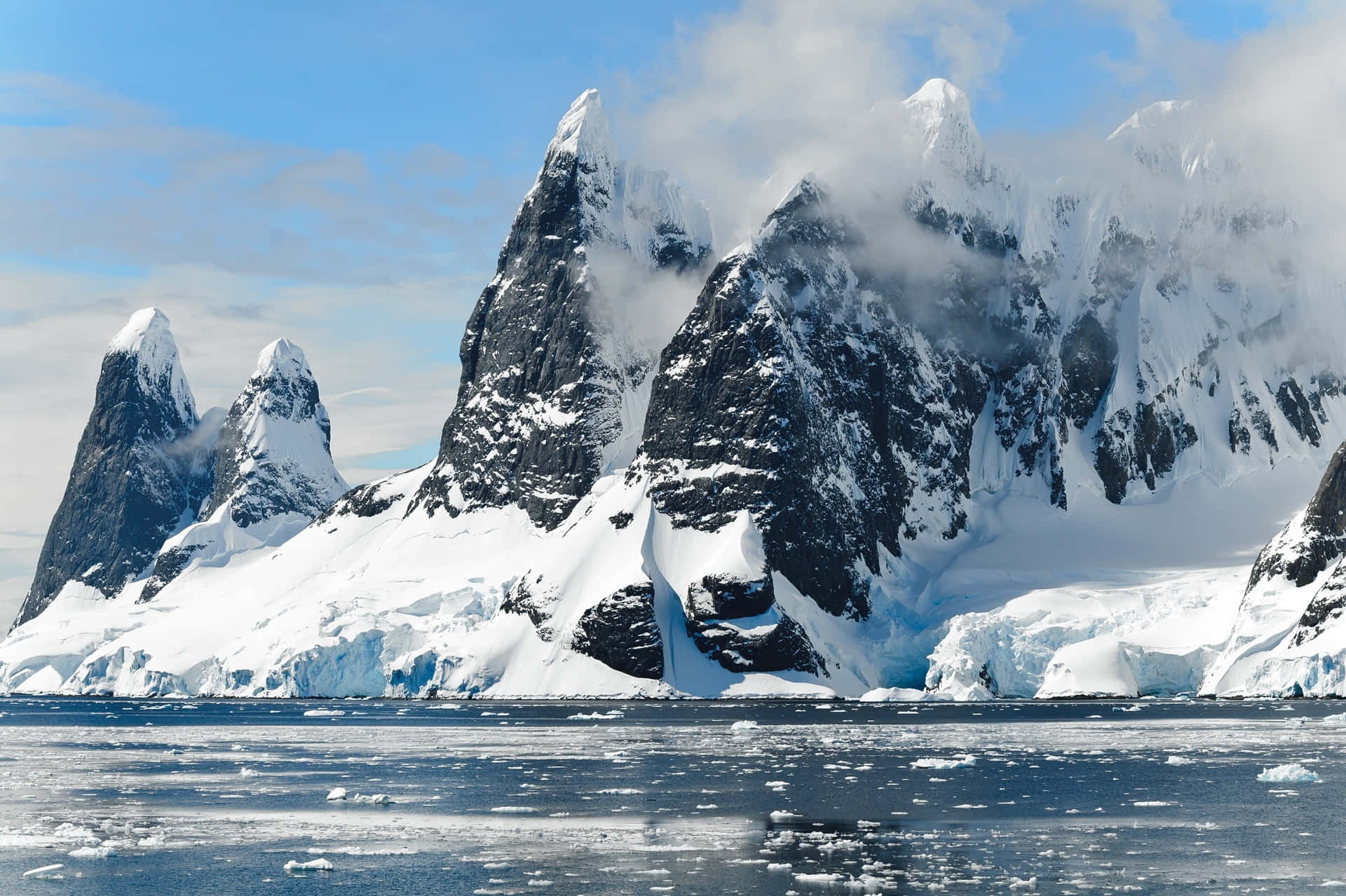 Antarctica 桌布 壁紙 — 白雪覆蓋的鋸齒狀山峰從冰冷的水域中升起，天空湛藍，飄著縷縷薄雲。