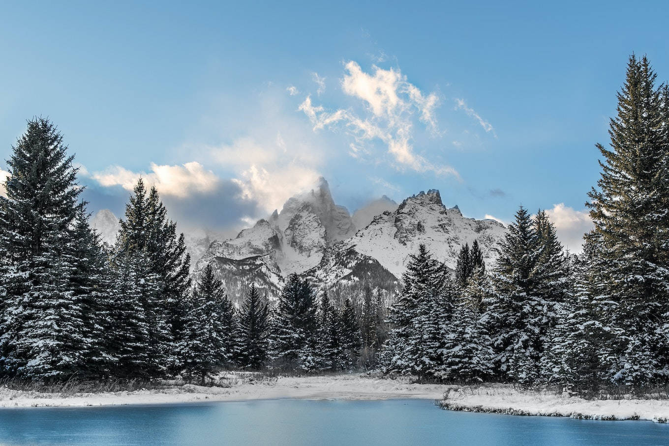 雪 桌布 壁紙 — 白雪覆蓋的鋸齒狀山峰聳立在冰凍的湖泊後面，周圍是灑滿白雪的常綠樹木，在明亮的藍天下飄著縷縷雲彩。