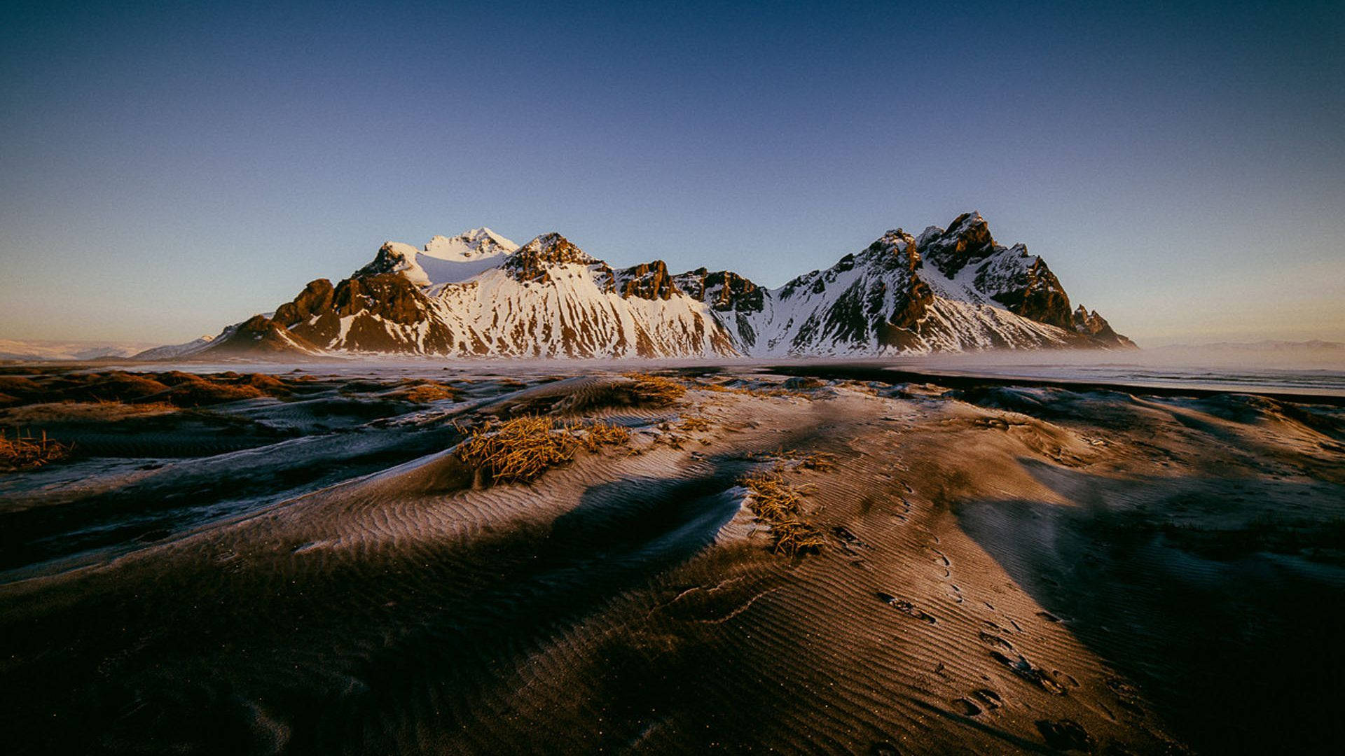 風景 桌布 壁紙 — 白雪覆蓋的鋸齒狀山脈在廣闊的黑沙灘後方升起，金色時刻中柔霧掠過沙丘。