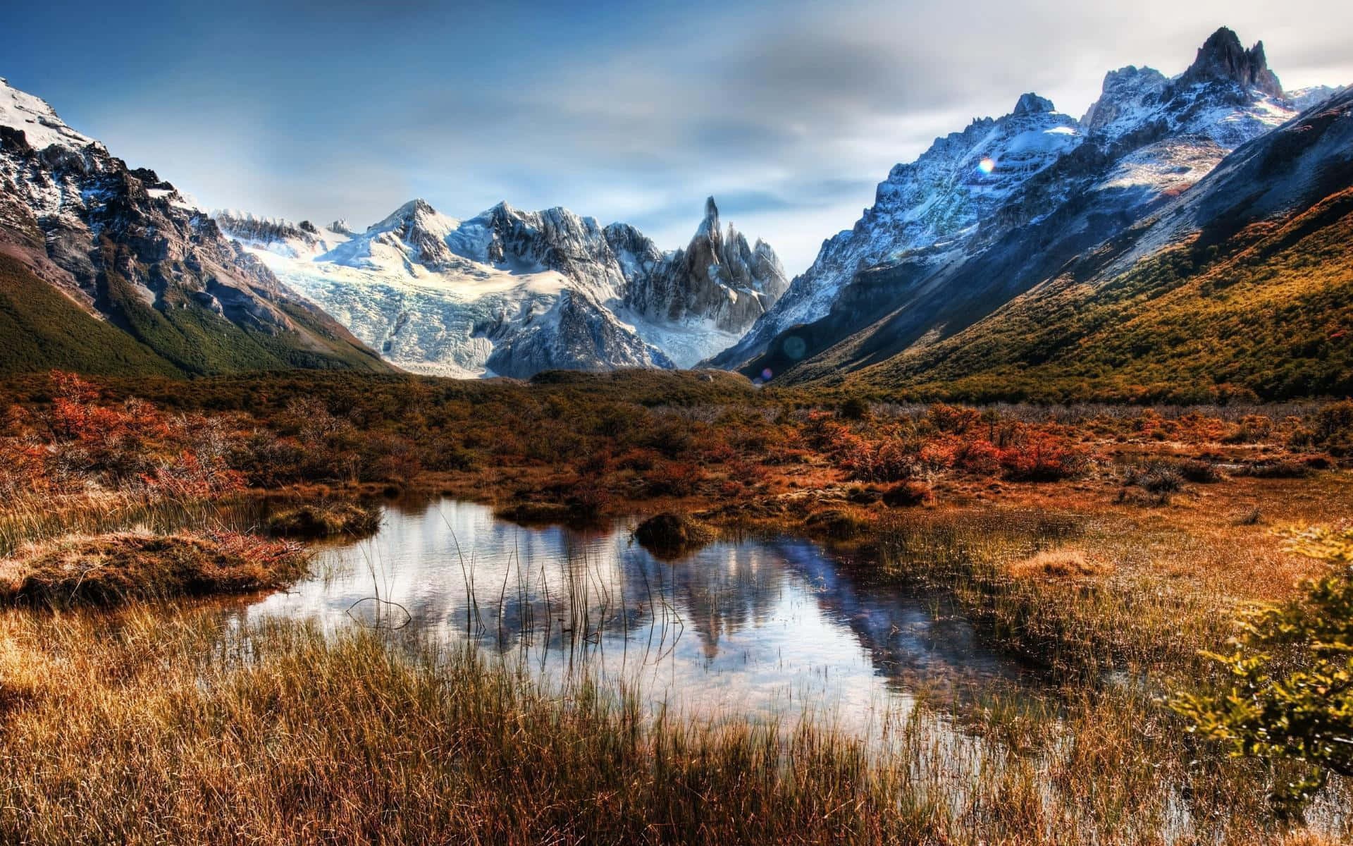 Patagonia 桌布 壁紙 — 白雪覆蓋的鋸齒狀山脈在部分多雲的天空下，從一個倒映著秋草的寧靜池塘後方升起。