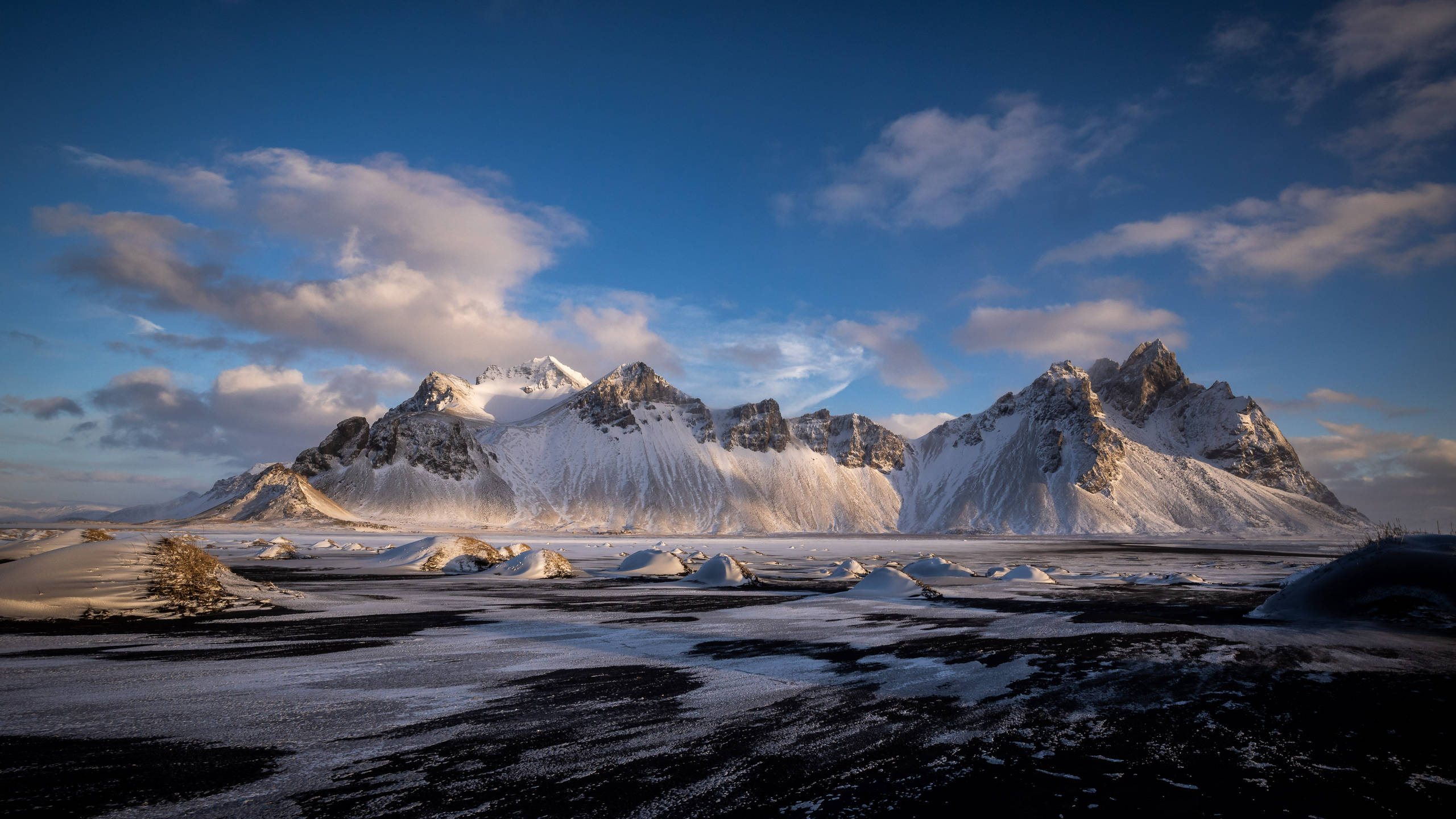 2560x1440 nature 桌布 壁紙 — 白雪覆蓋的鋸齒狀山脈在金色時刻戲劇性地聳立在點綴著散雲的鮮豔藍天下。