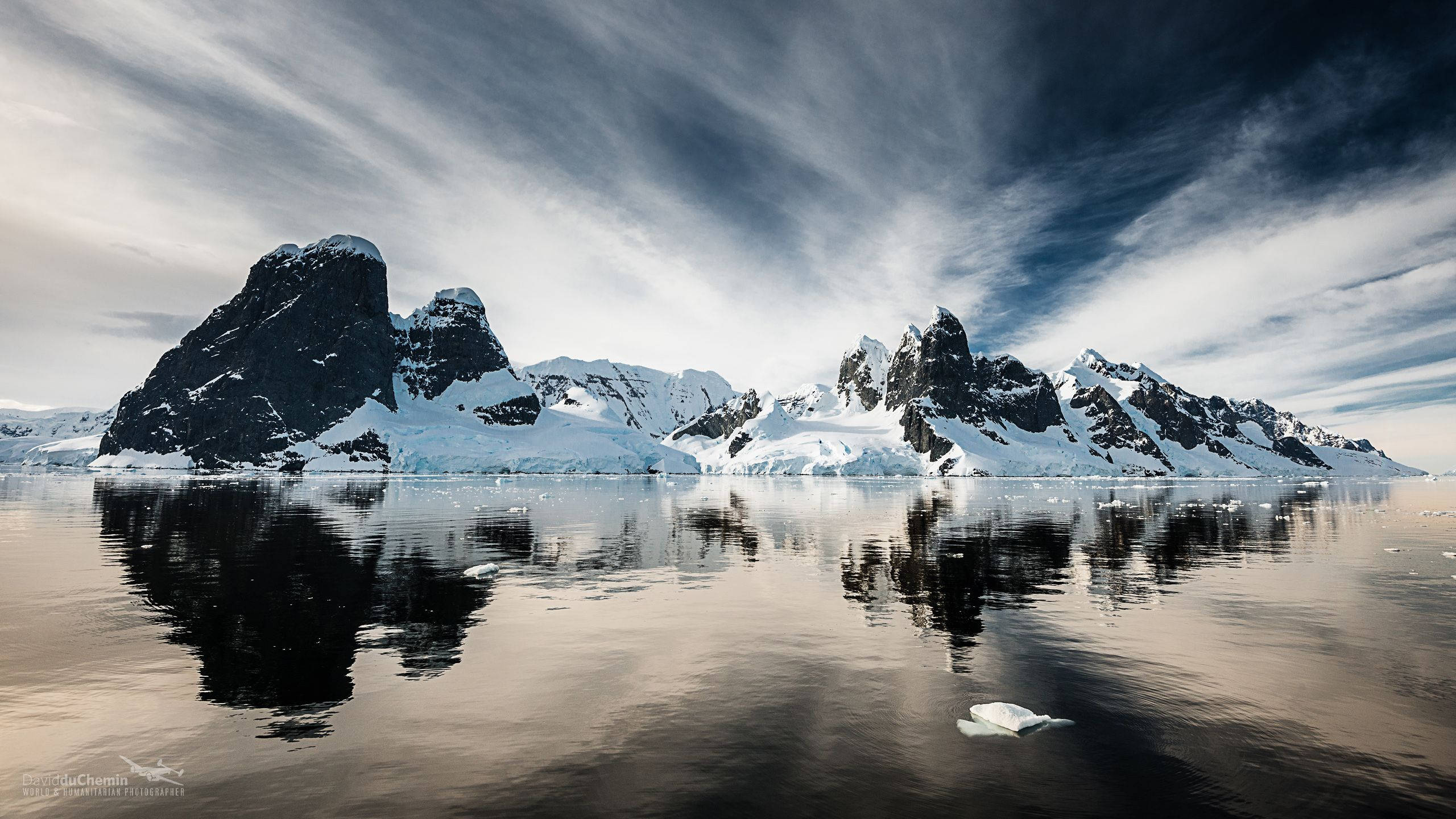 Antarctica 桌布 壁紙 — 白雪覆蓋的鋸齒狀山脈從平靜的水面升起，在戲劇性的多雲天空下呈現完美的倒影。