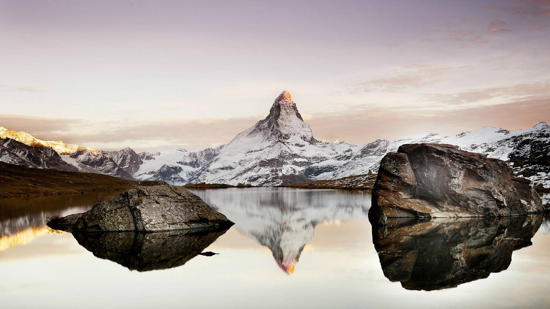 Alps 桌布 壁紙 — 白雪覆蓋的馬特洪峰在日出時發光，完美倒映在一個由大石圍繞的平靜湖面。