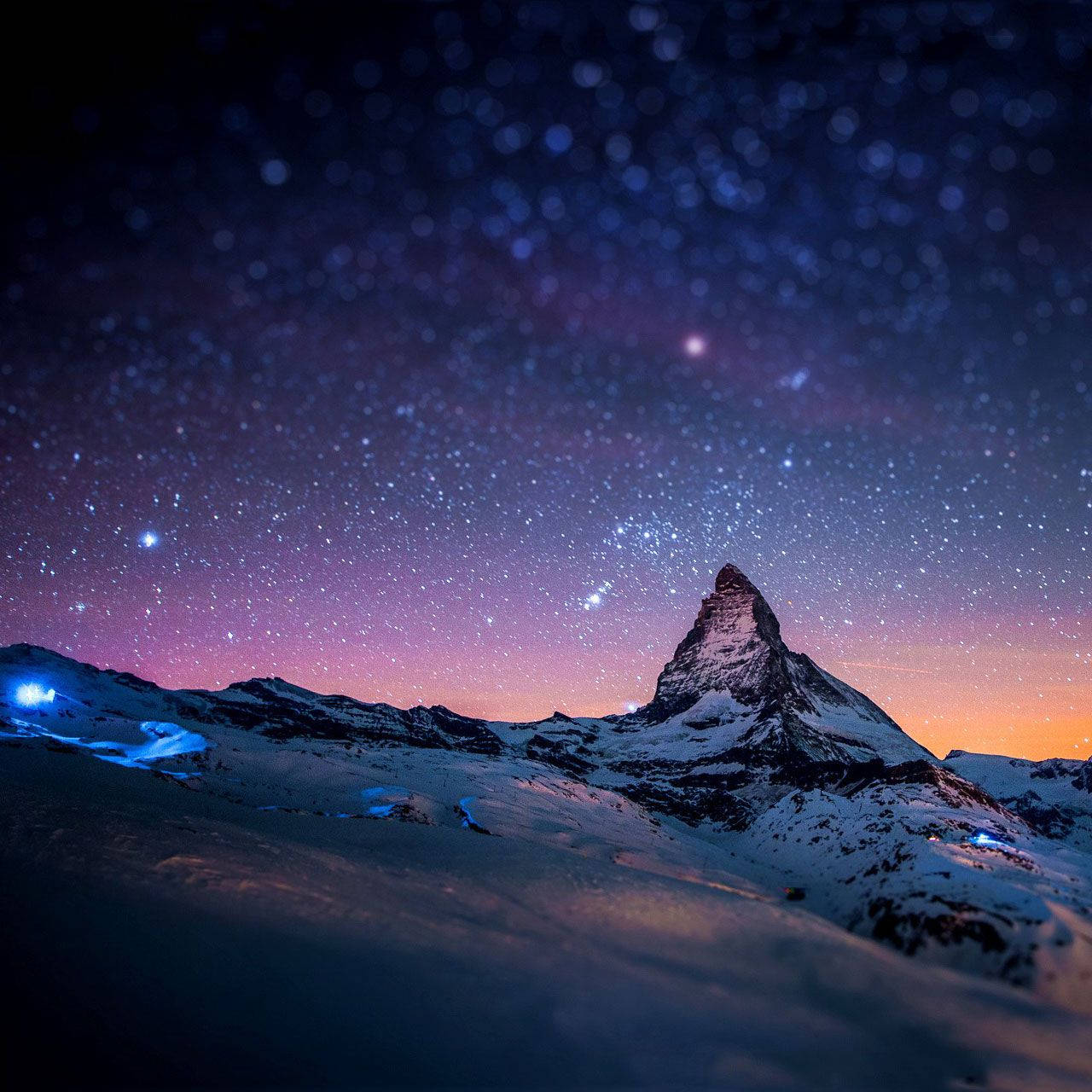 夜空 桌布 壁紙 — 白雪覆蓋的馬特洪峰在星光燦爛的暮色天空下聳立，帶有紫色和橙色色調，山坡上還有發光的藍色燈光。