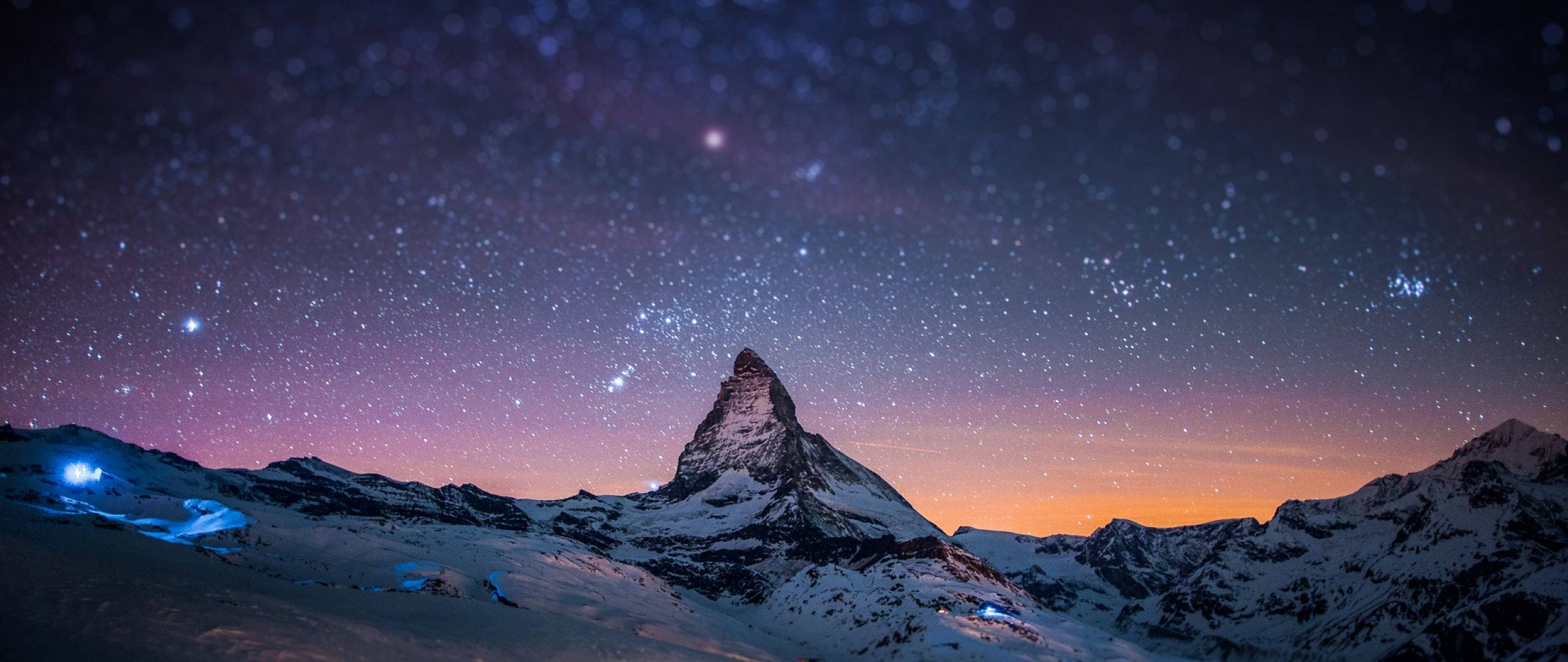 山景 桌布 壁紙 — 白雪覆蓋的馬特洪峰在星空暮色下升起，紫橙色地平線和下方發光的藍色燈光。