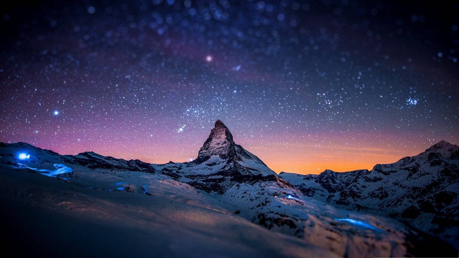 美麗高畫質 桌布 壁紙 — 白雪覆蓋的馬特洪峰在星空暮色下聳立，帶有紫橙色漸變和下方發光的藍色燈光。