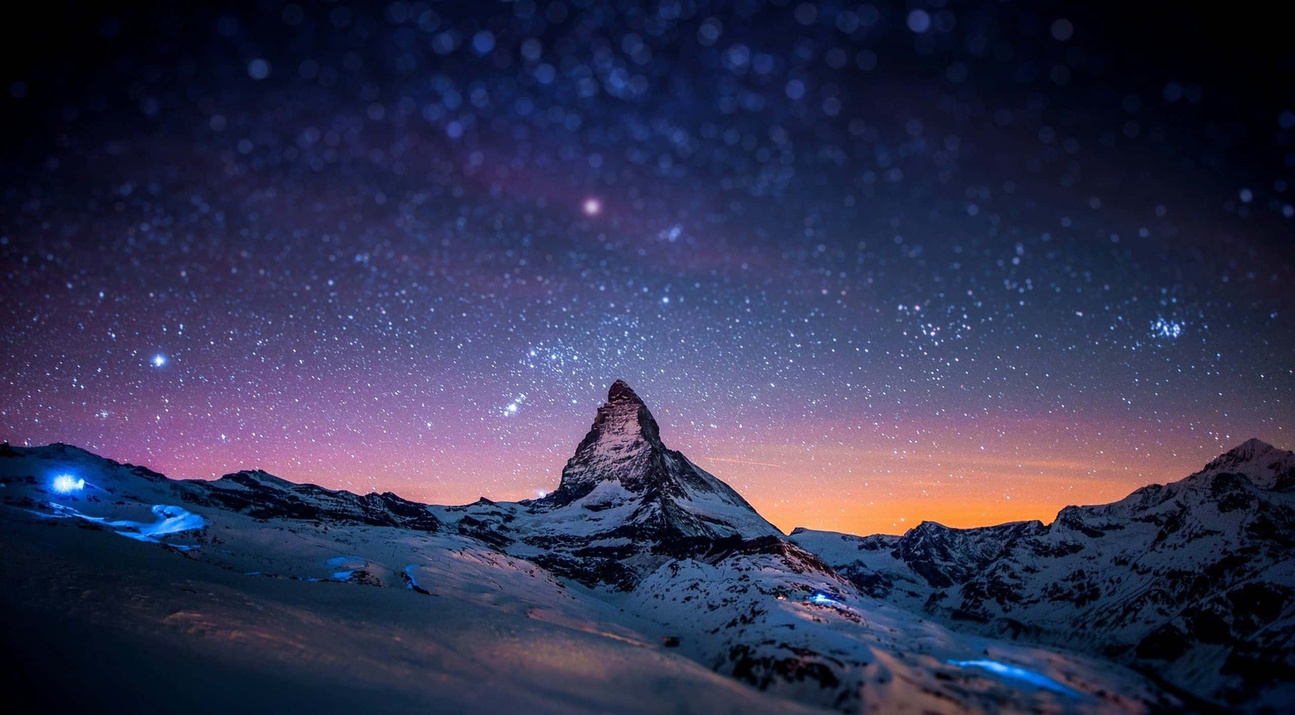 山景 桌布 壁紙 — 白雪覆蓋的馬特洪峰在星空暮色天空的襯托下形成剪影，天空帶有紫橙色漸變，下方有發光的藍色燈光。