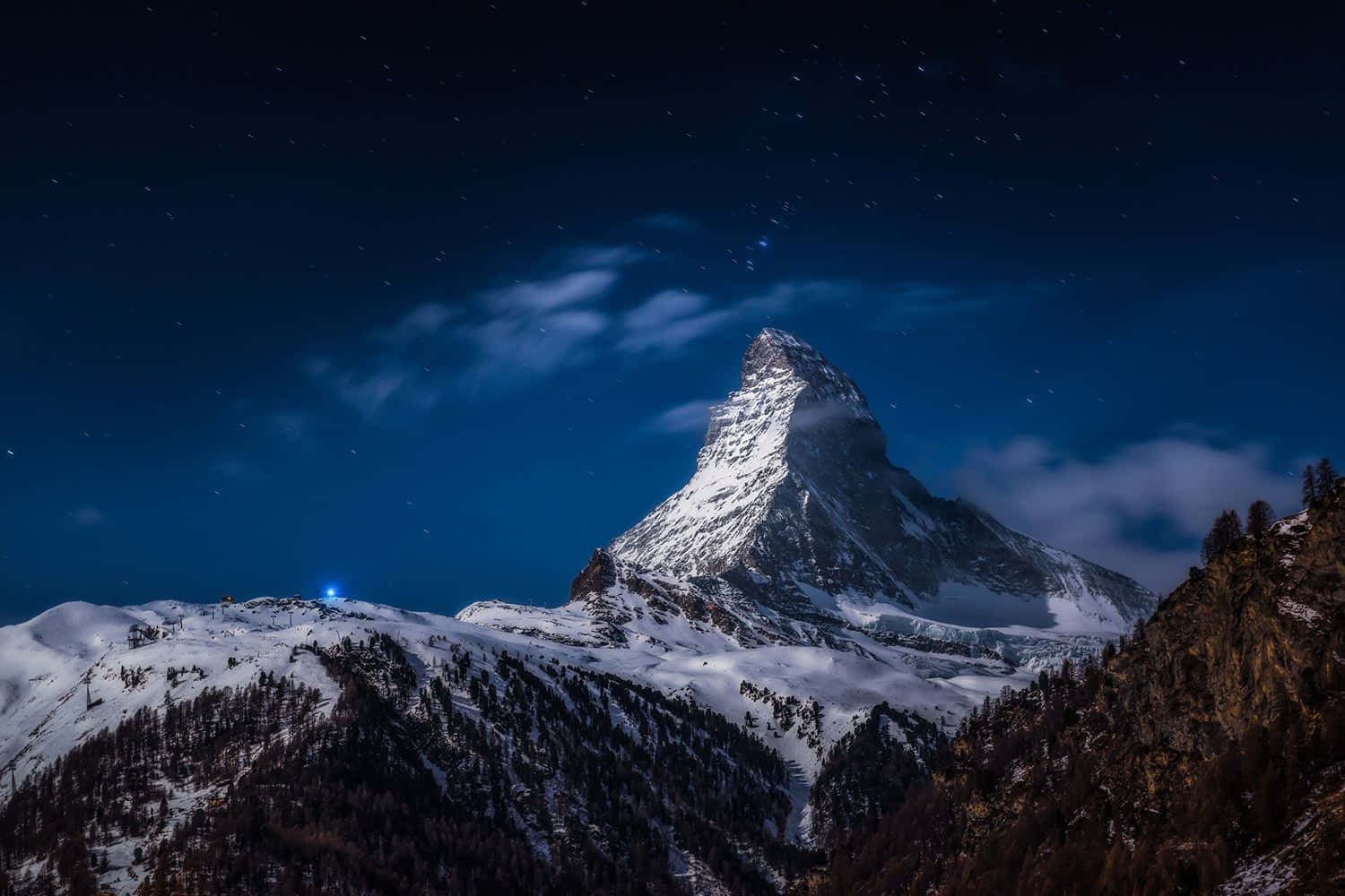Night Aesthetic 桌布 壁紙 — 白雪覆蓋的馬特洪峰在繁星點點的夜空下巍峨聳立，伴隨著飄動的雲朵和下方的一盞藍光。