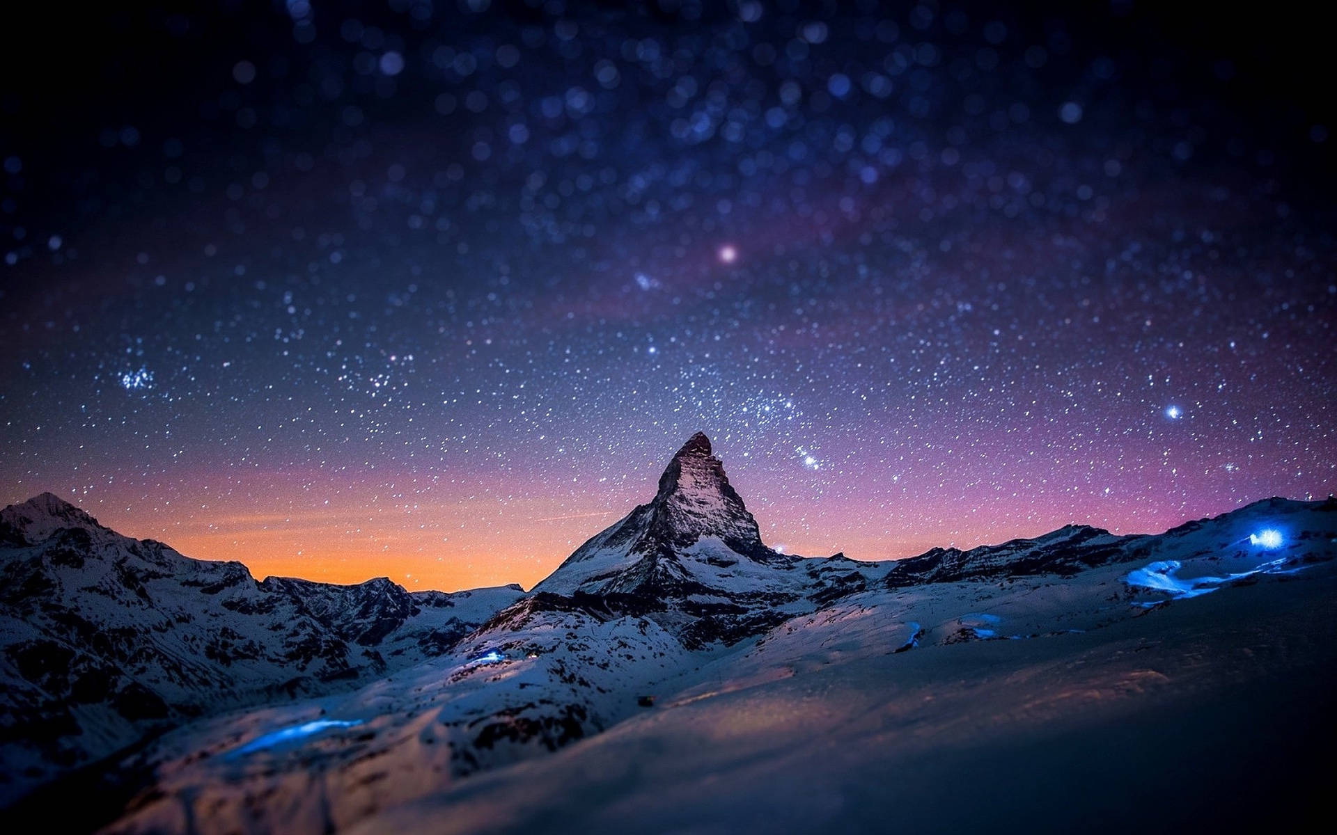 Alps 桌布 壁紙 — 白雪覆蓋的馬特洪峰在繁星點點的暮色天空下聳立，地平線呈現紫橙色，下方有發光的藍色燈光。