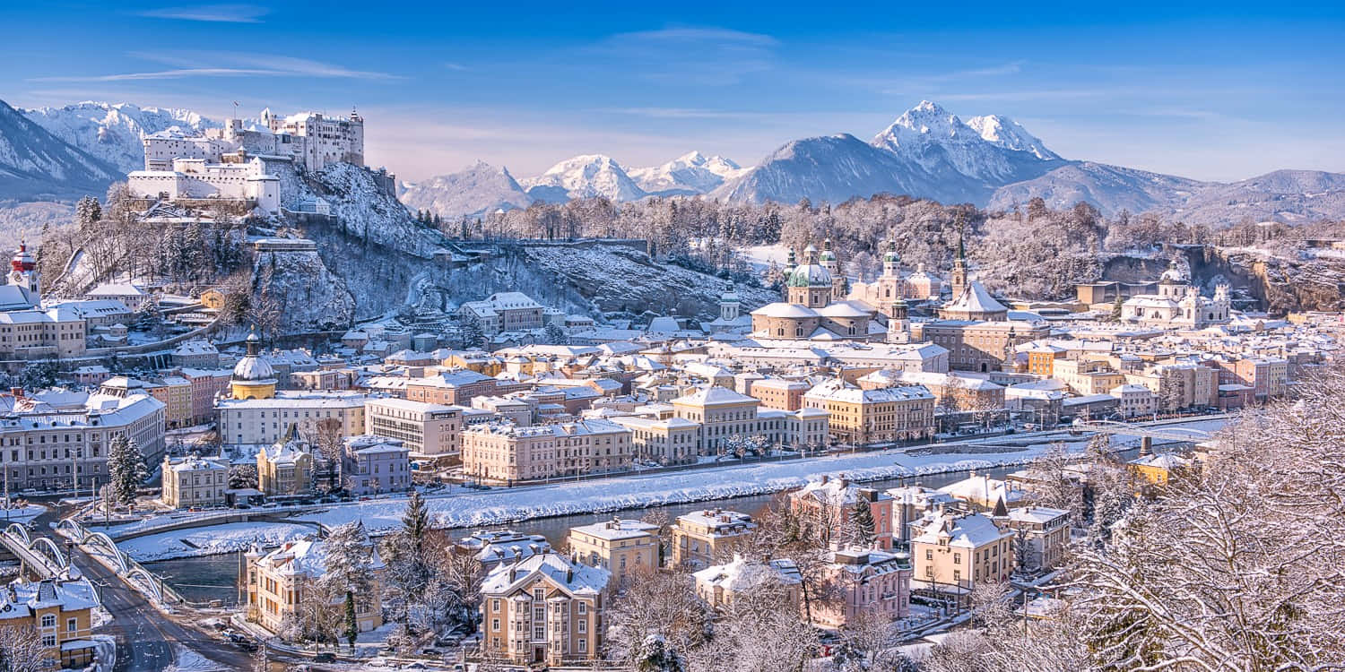 雪 桌布 壁紙 — 白雪覆蓋著一個坐落在山谷中的歷史歐洲城市，背後是白雪覆蓋的山脈，在清澈的藍天下聳立。