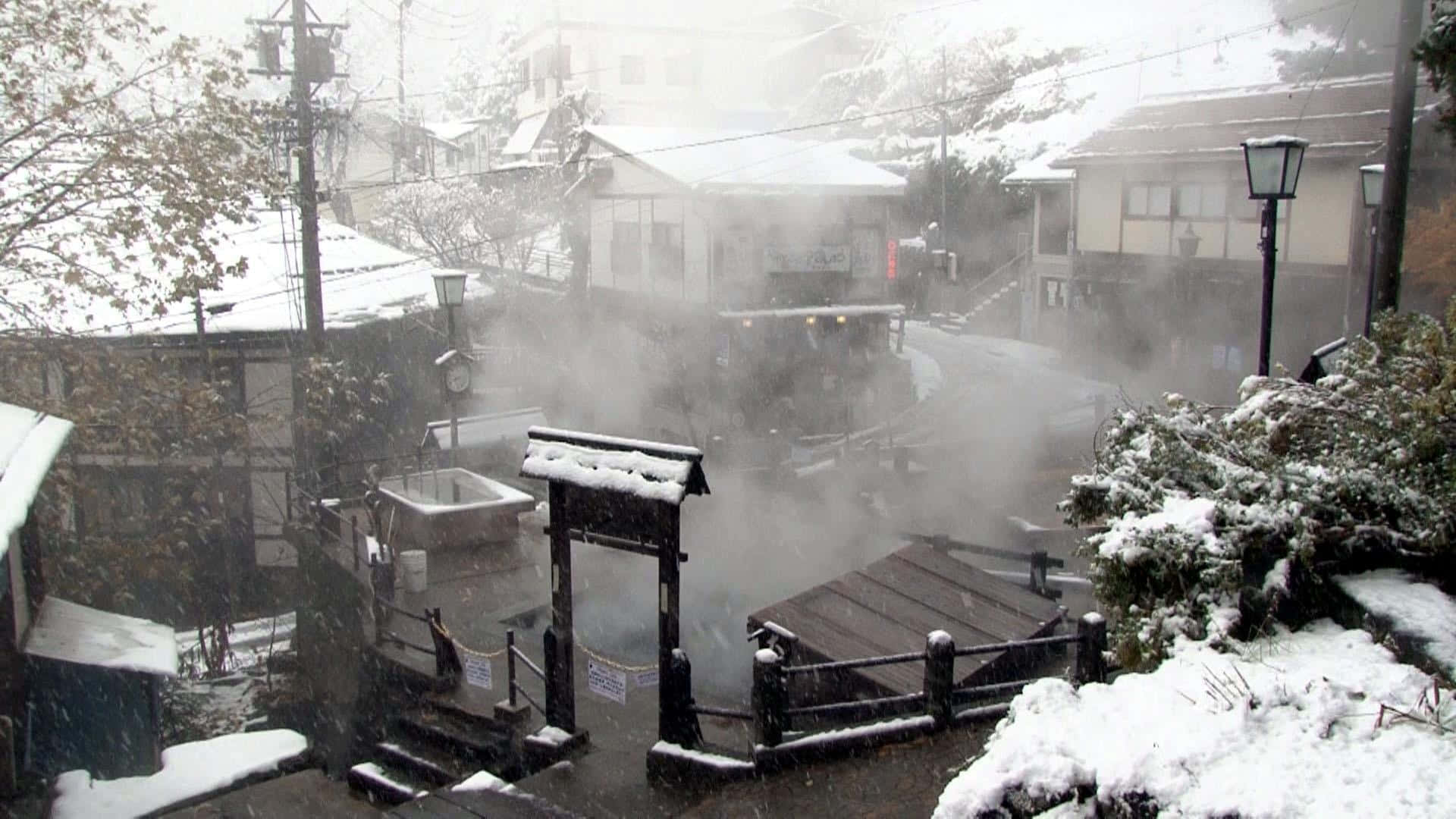 High Resolution Snow 桌布 壁紙 — 白雪覆蓋著一個寧靜的日本溫泉村莊，蒸汽從溫泉浴池中升起，伴隨著飄落的雪花。
