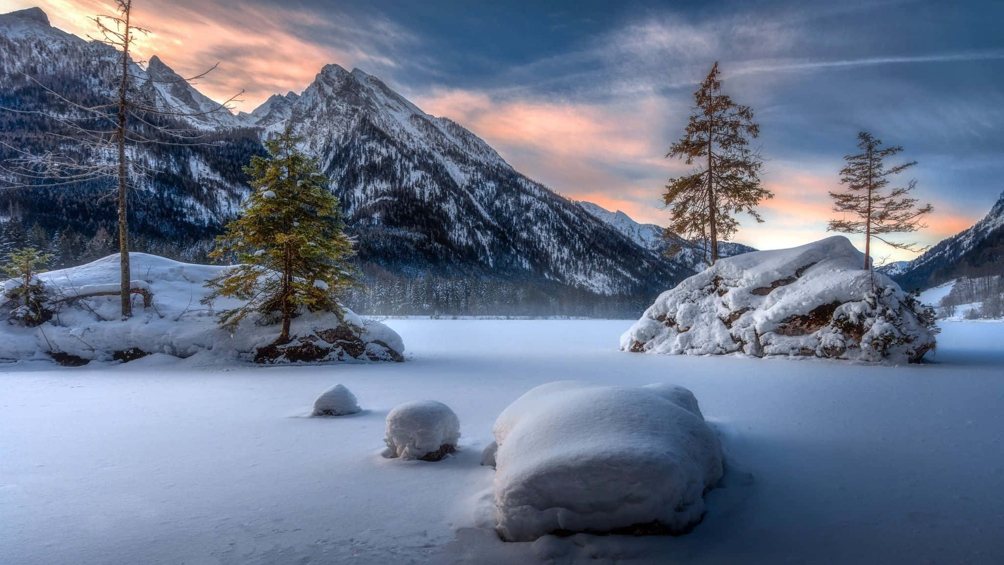 Snowy Landscape 桌布 壁紙 — 白雪覆蓋著一個被高聳山脈環繞的冰凍湖泊，在塗上柔和粉紅和藍色調的暮色天空下。