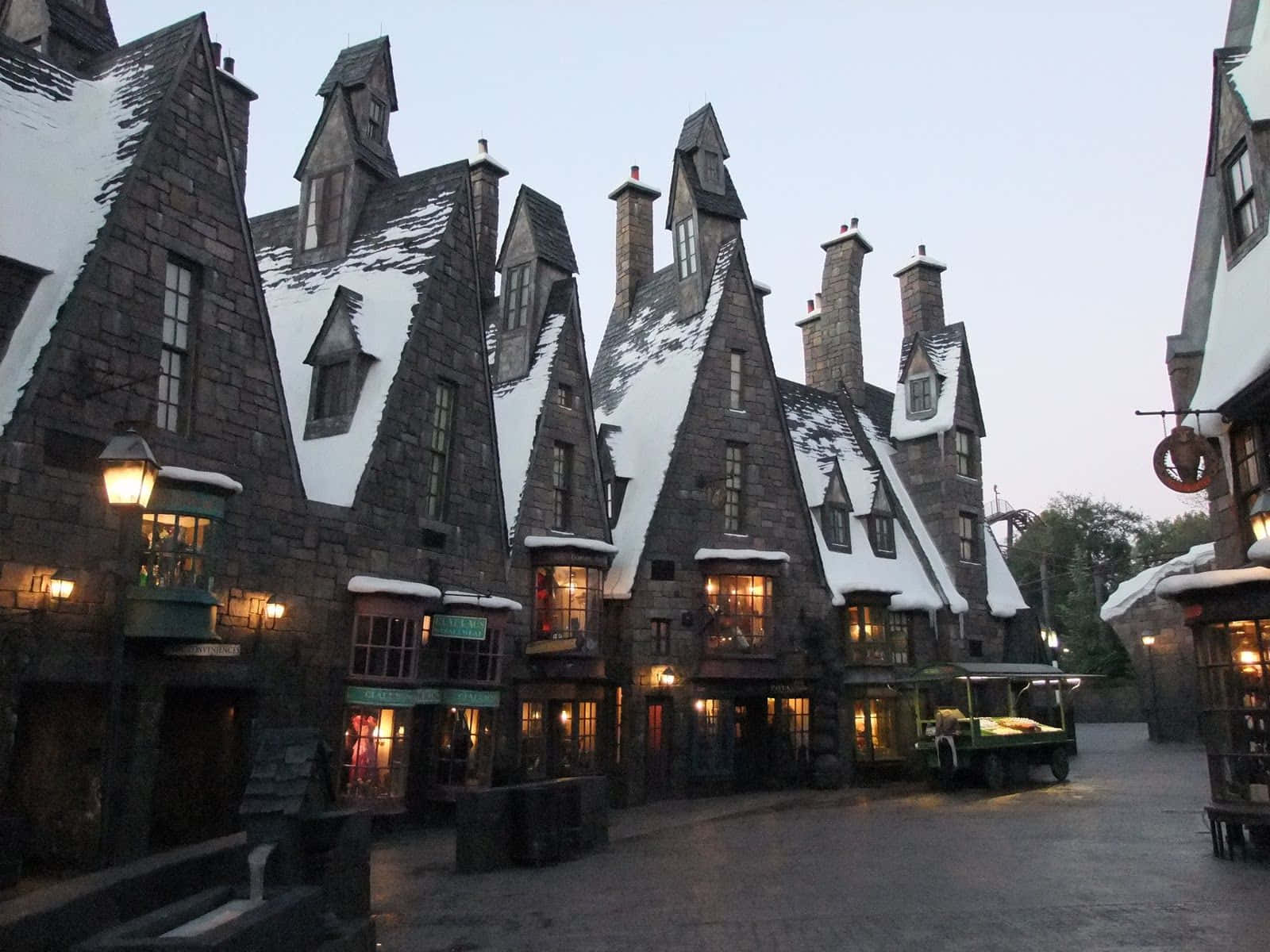 Hogsmeade 桌布 壁紙 — 白雪覆蓋著中世紀風格石砌建築的陡峭山牆屋頂，溫暖的燈光從窗戶中透出，映照在黃昏時分寧靜的村莊街道上。