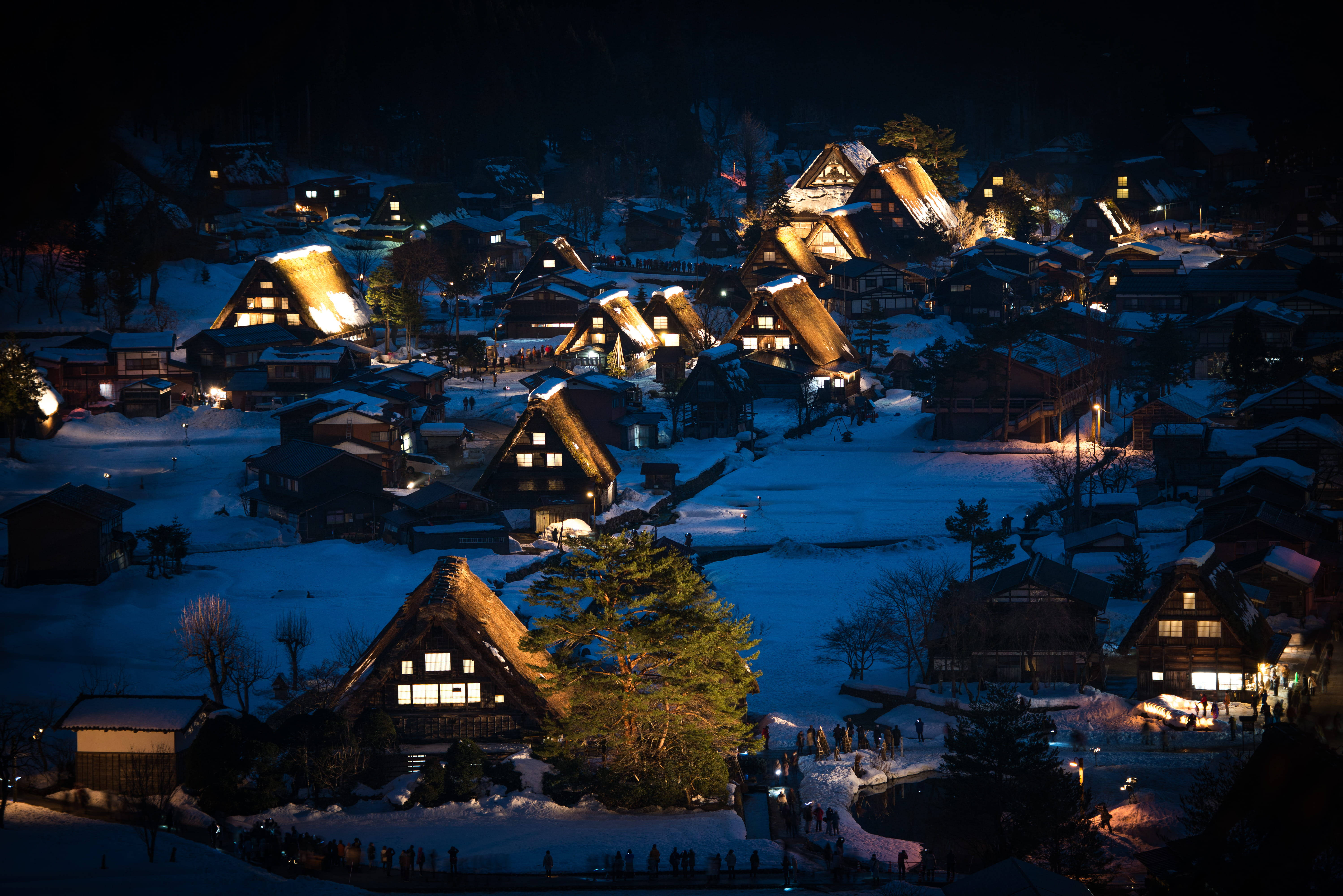 日本 桌布 壁紙 — 白雪覆蓋著傳統茅草屋頂的房屋，山間村莊在夜空下溫暖地發光。