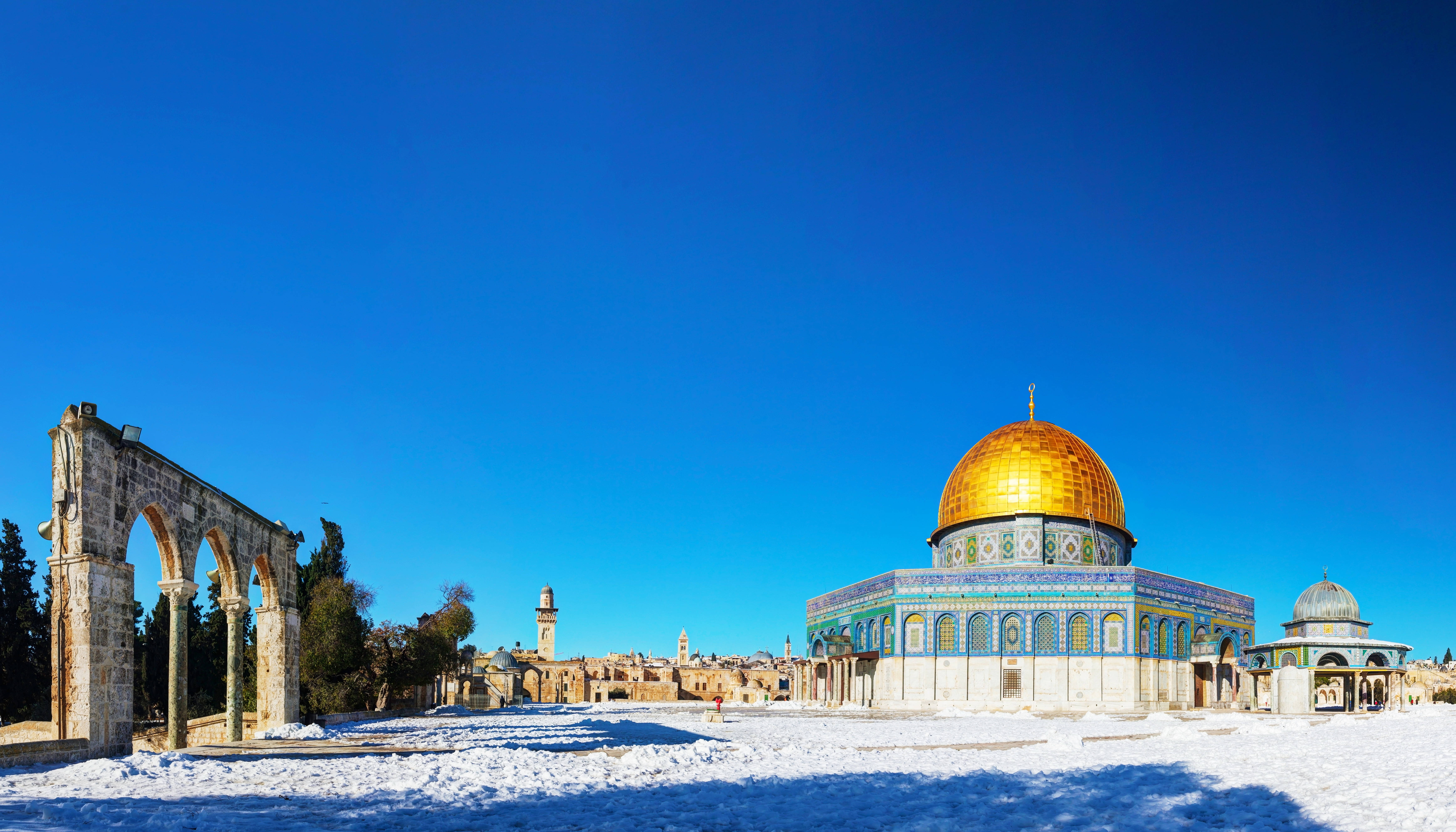 muslim 桌布 壁紙 — 白雪覆蓋著古老的石拱門和金色的圓頂清真寺，天空湛藍清澈。