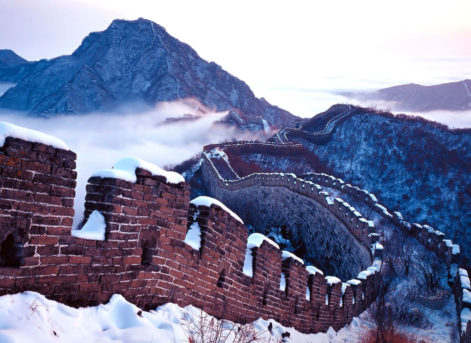 中國 桌布 壁紙 — 白雪覆蓋著古老的長城，在柔和的黎明天空下蜿蜒穿過霧氣繚繞的山巒。