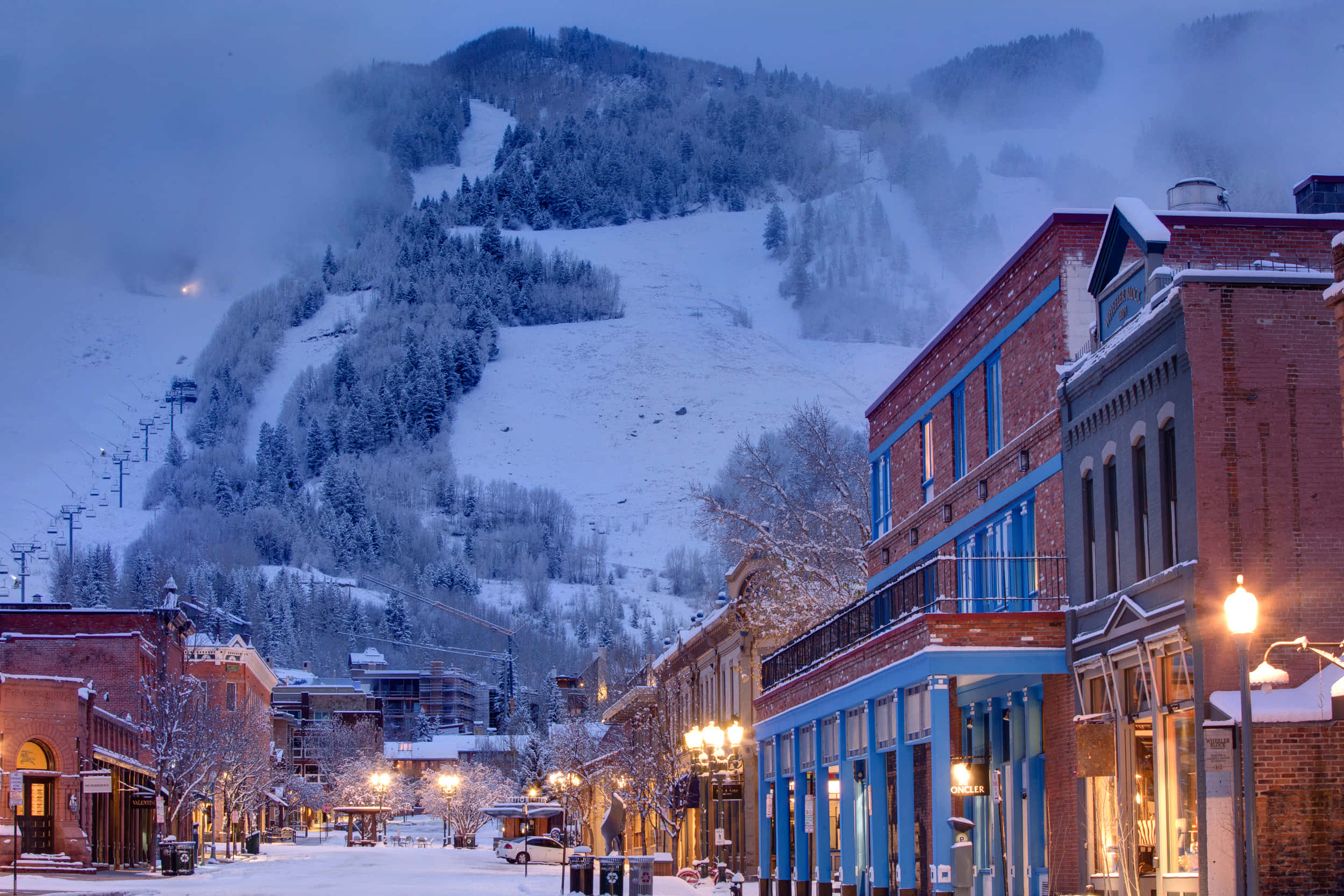 Colorado 桌布 壁紙 — 白雪覆蓋著寧靜的山城街道，在暮色中，發光的街燈和後方霧氣繚繞的山坡上的滑雪纜車清晰可見。