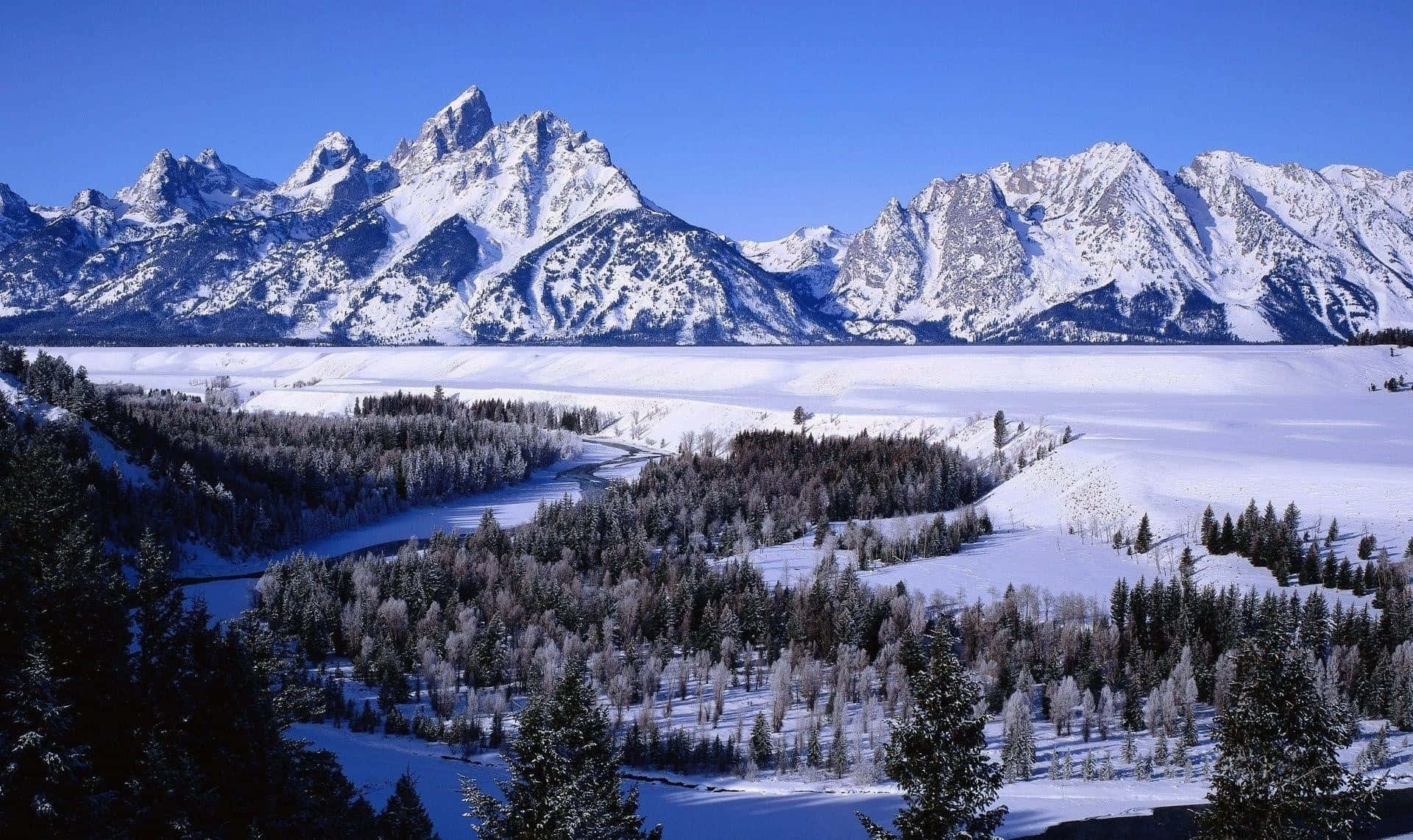 山景 桌布 壁紙 — 白雪覆蓋著寧靜的山谷，有常綠樹和蜿蜒的河流，在清澈的藍天下聳立著參差不齊的山峰。