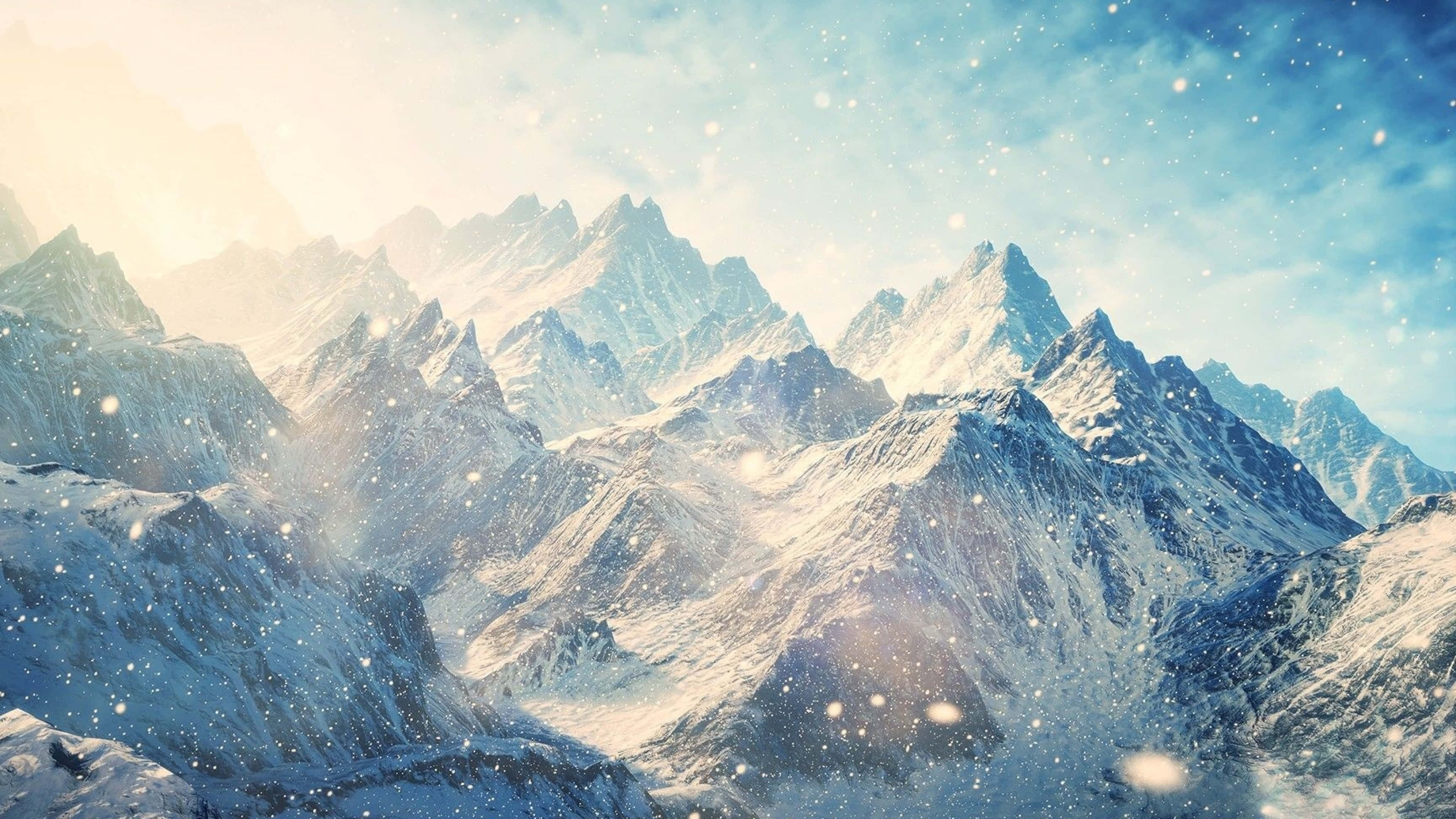 Skyrim Landscape 桌布 壁紙 — 白雪覆蓋著崎嶇的山峰，在柔和的藍天下，飄落的雪花捕捉著金色的陽光。