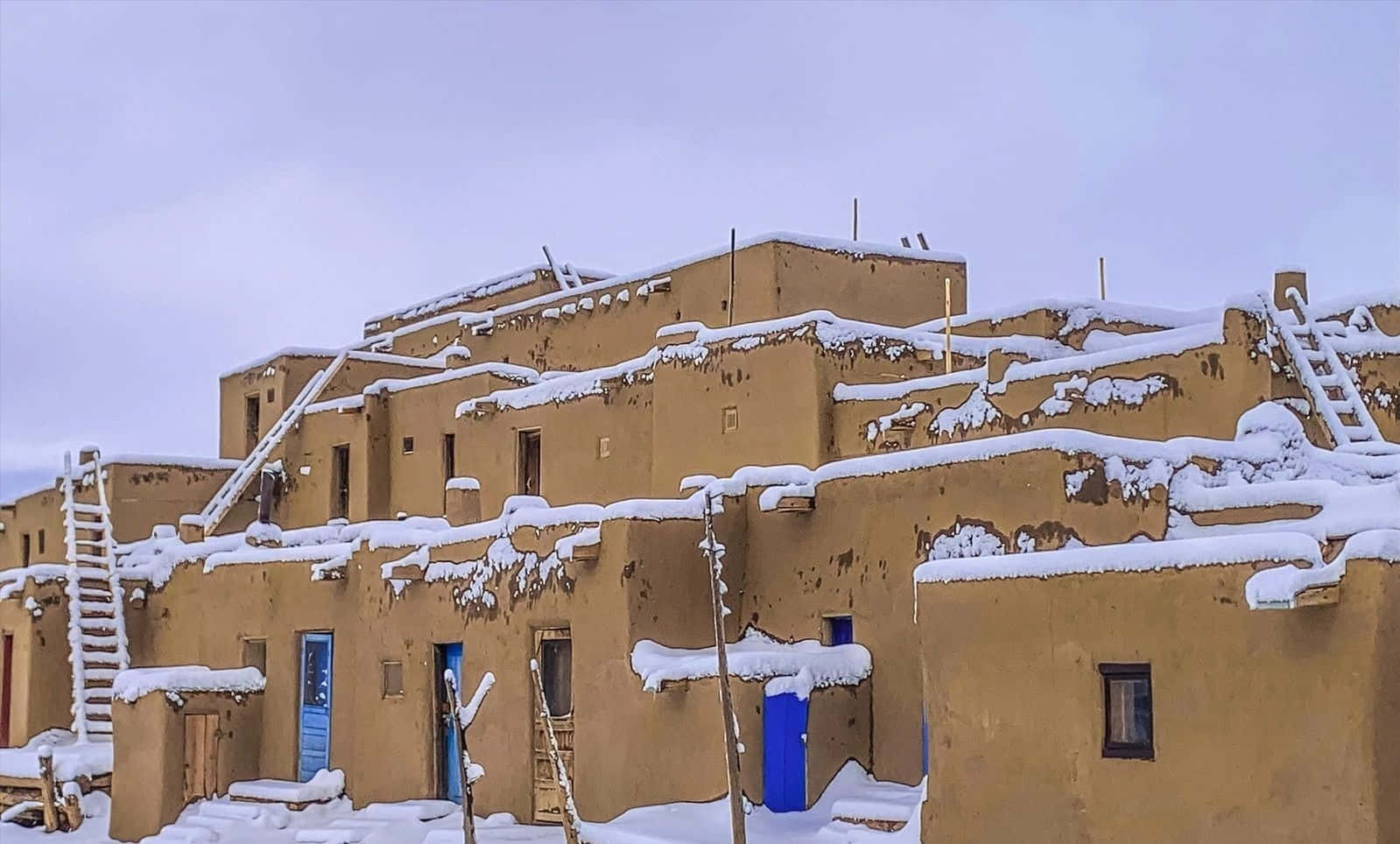 Cool Places 桌布 壁紙 — 白雪覆蓋著藍色門的土坯建築，在蒼白的冬日天空下。