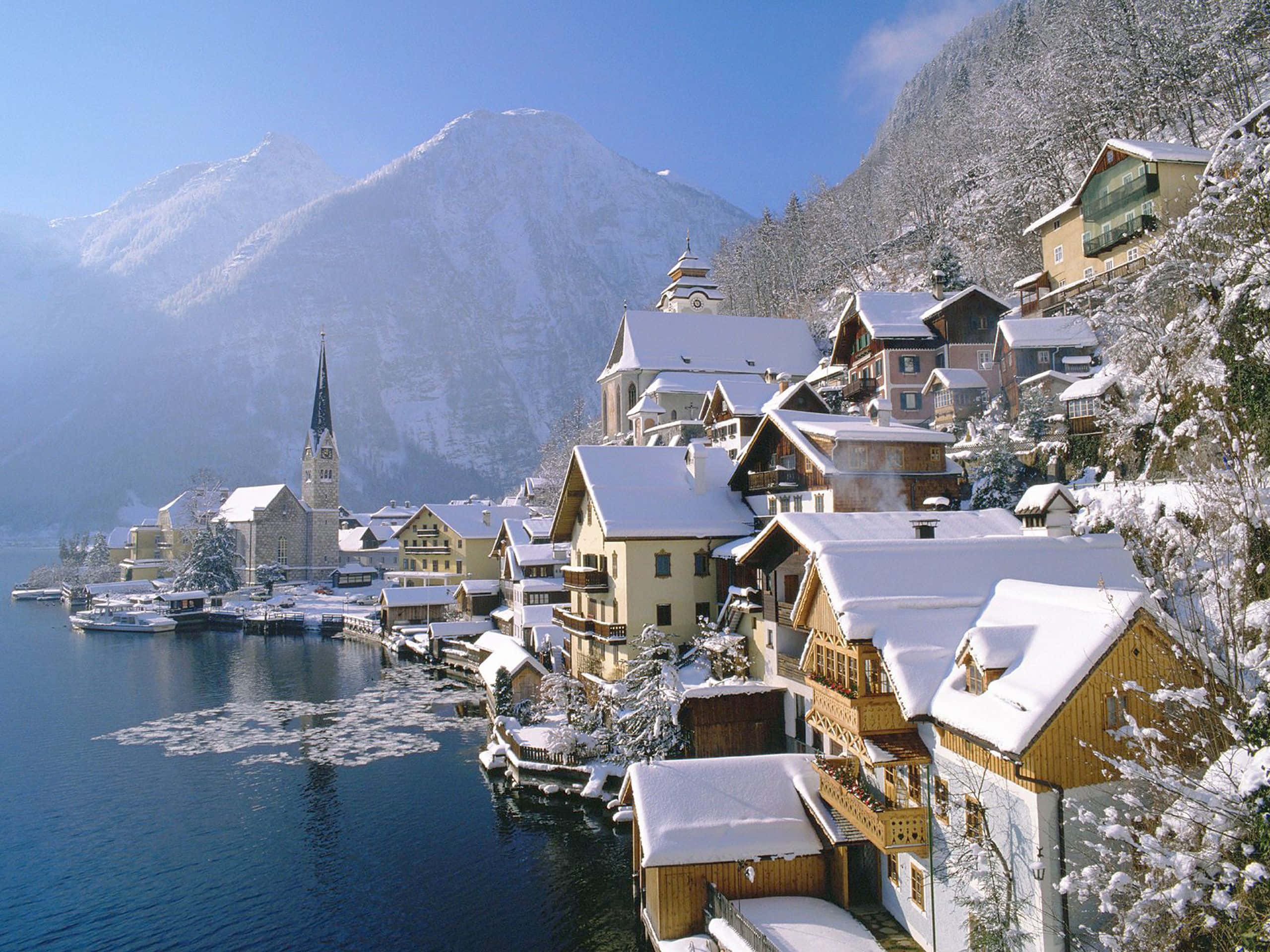 Snowy Village 桌布 壁紙 — 白雪覆蓋著迷人的阿爾卑斯山房屋，擁抱著寧靜的湖泊，雄偉的山脈在清澈的藍天下聳立於後。