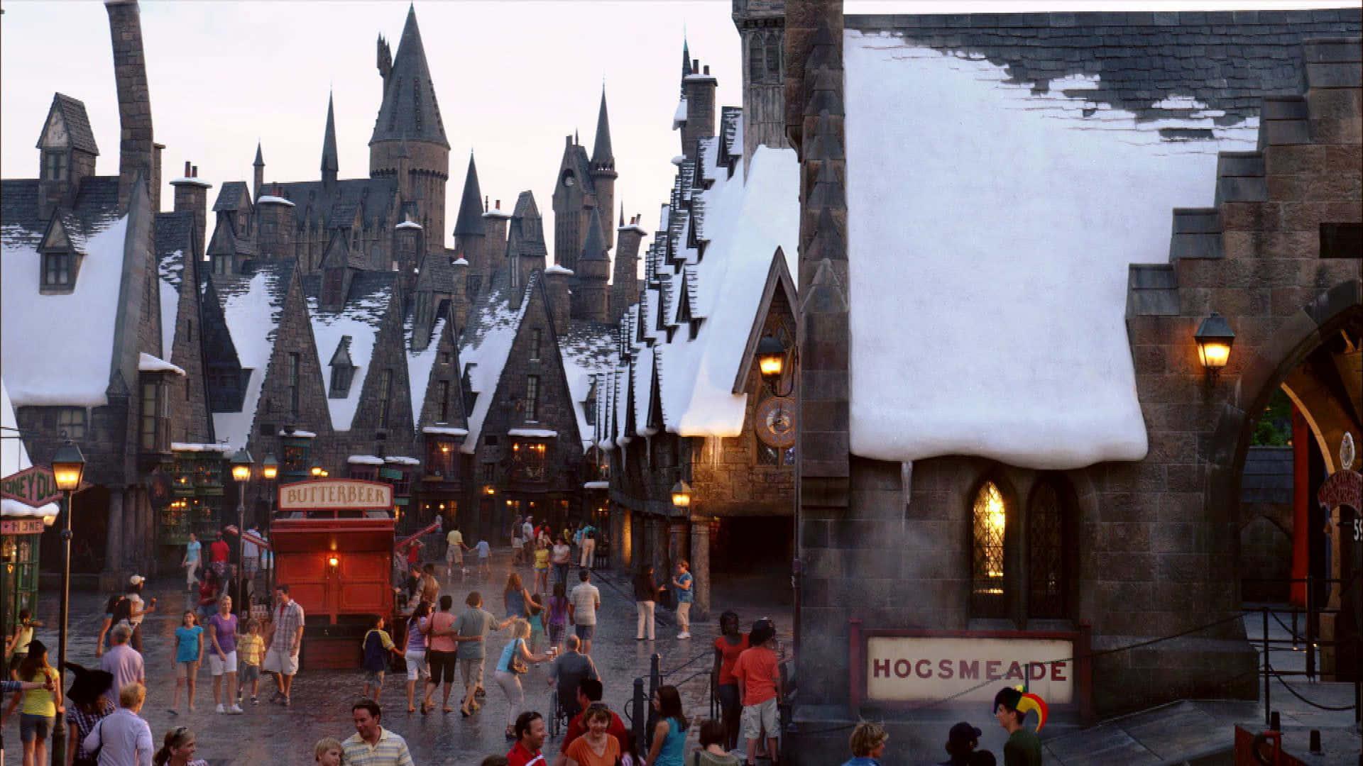 Hogsmeade 桌布 壁紙 — 白雪覆蓋著霍格莫德村的屋頂，遊客在黃昏時分漫步於溫暖燈光照亮的鵝卵石街道上。