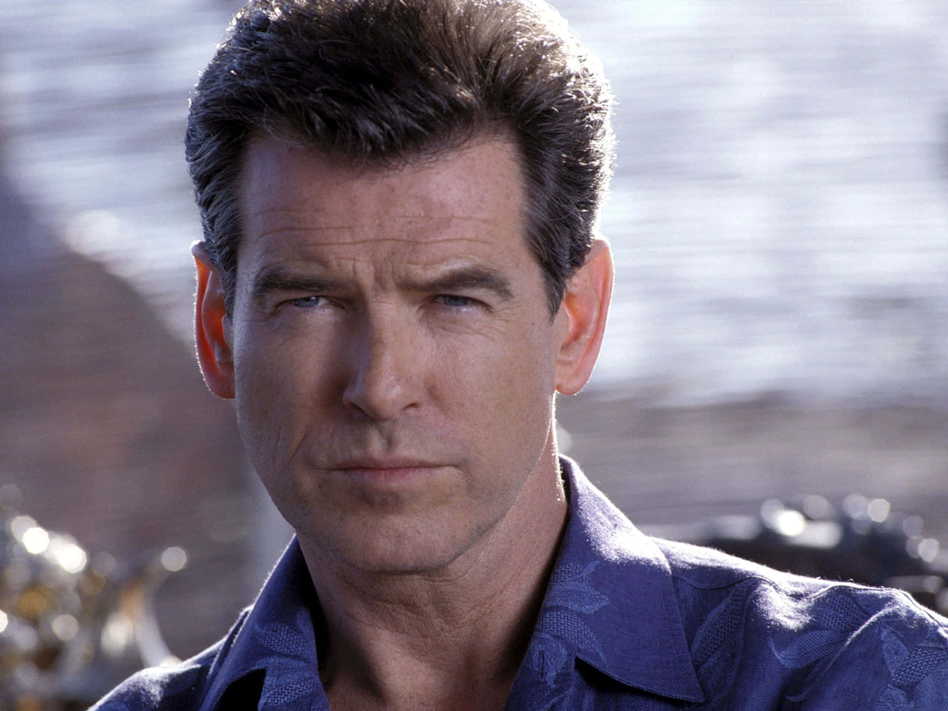 pierce brosnan 桌布 壁紙 — 皮爾斯·布洛斯南的特寫，擁有強烈的藍色眼睛和深色頭髮，穿著藍色圖案襯衫，背景是模糊的水面。
