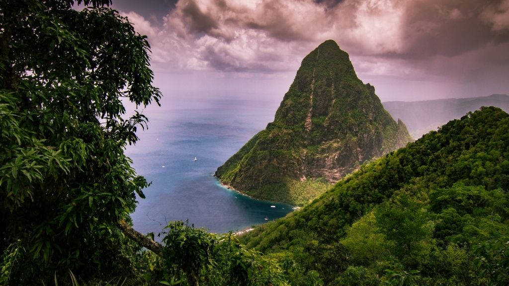山脈 桌布 壁紙 — 皮通山從鬱鬱蔥蔥的綠色山坡聳立到綠松石水域上方陰鬱的紫色天空的戲劇性景色。