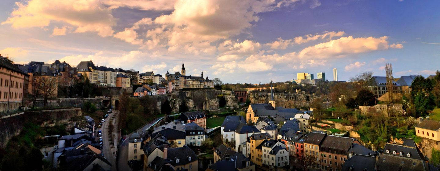 Luxembourg 桌布 壁紙 — 盧森堡市日落全景，歷史建築坐落於岩石地形，遠處有現代摩天大樓。