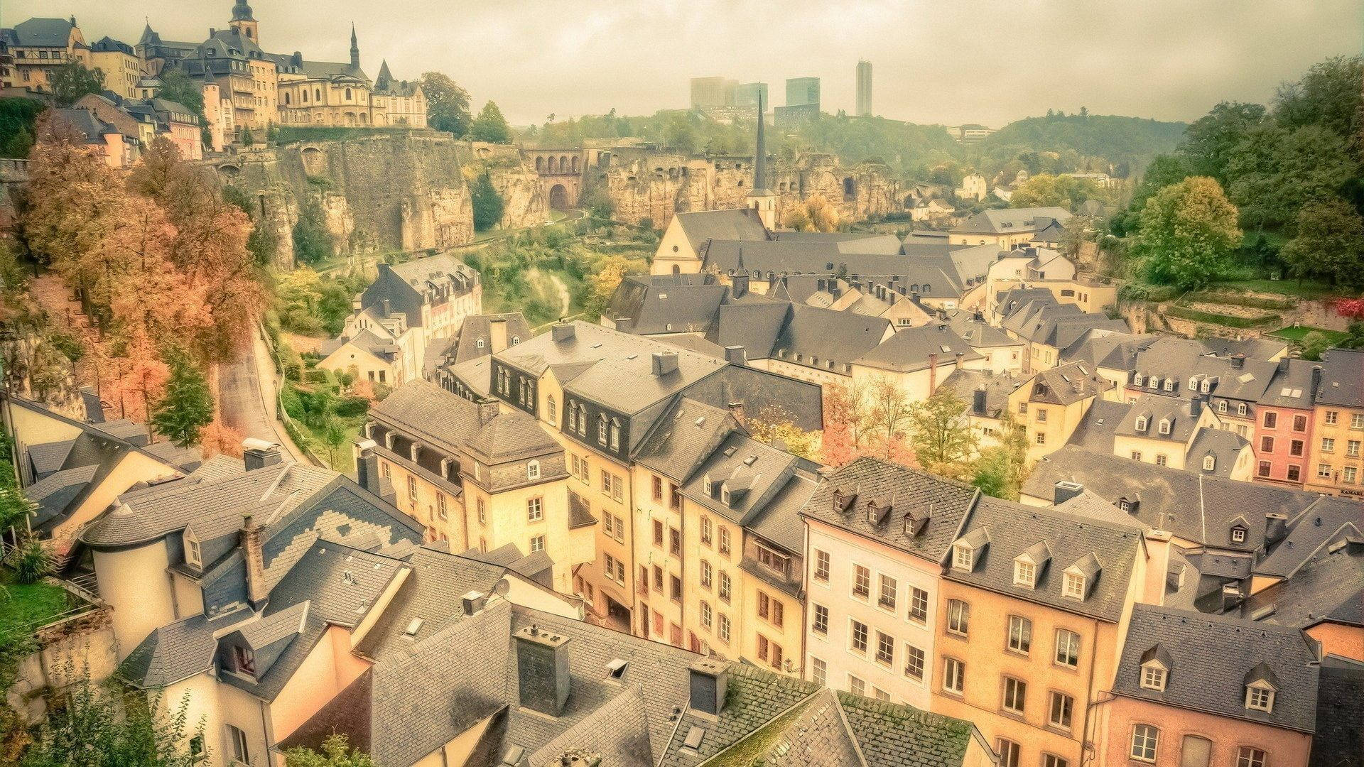 Luxembourg 桌布 壁紙 — 盧森堡市的空拍圖，歷史建築坐落在山坡上，沐浴在柔和的金色光線中。