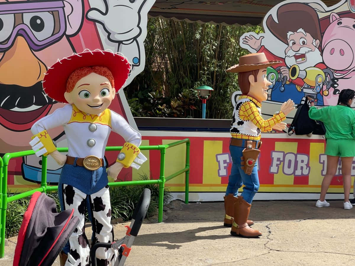 玩具總動員 桌布 壁紙 — 真人大小嘅《玩具總動員》角色 Jessie 同 Woody 喺明亮日光下嘅一個充滿活力嘅主題公園景點歡迎賓客。