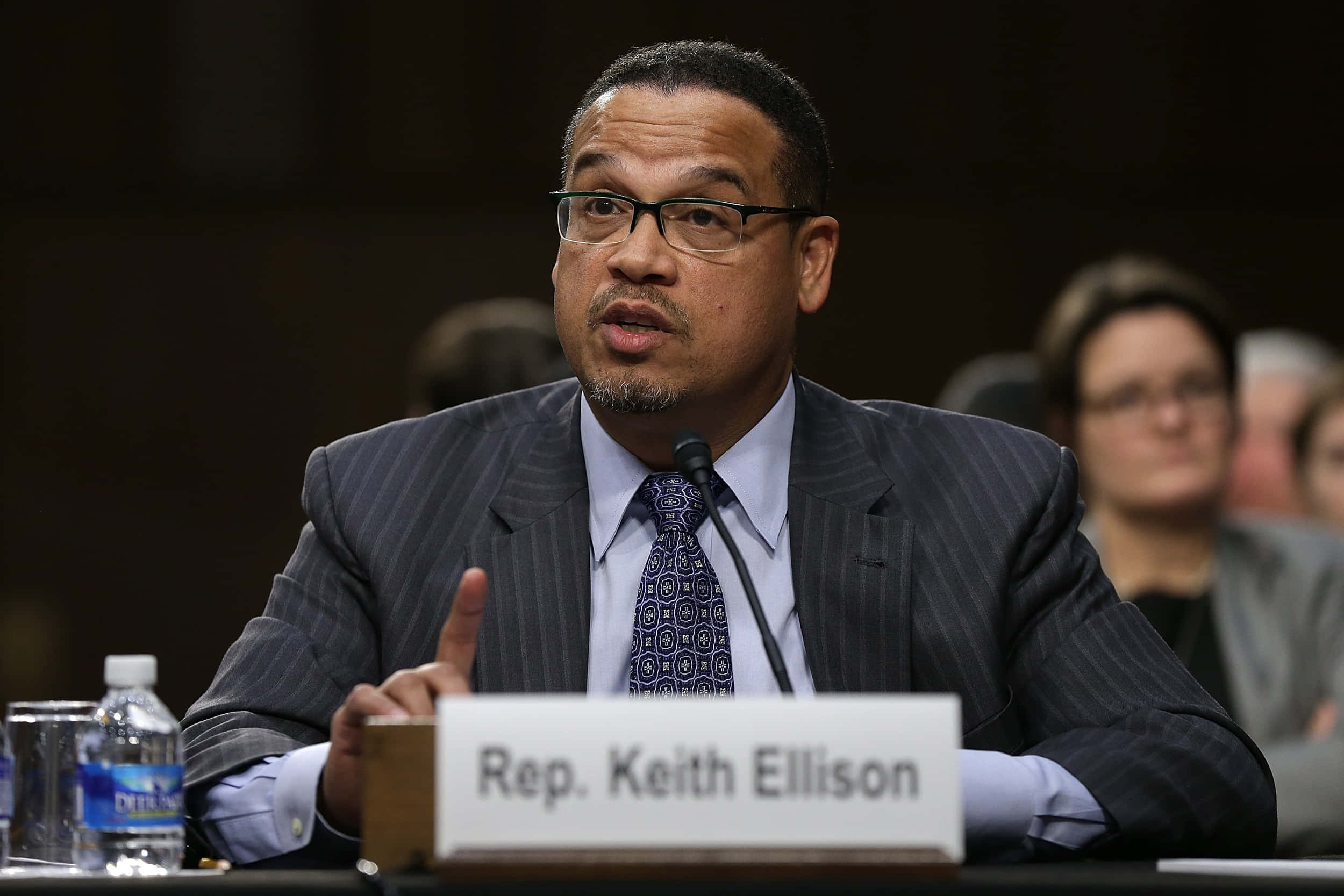 Famous 桌布 壁紙 — 眾議員 Keith Ellison 在正式聽證會上發言，身穿細條紋西裝和圖案領帶，桌上放著名牌和水瓶。