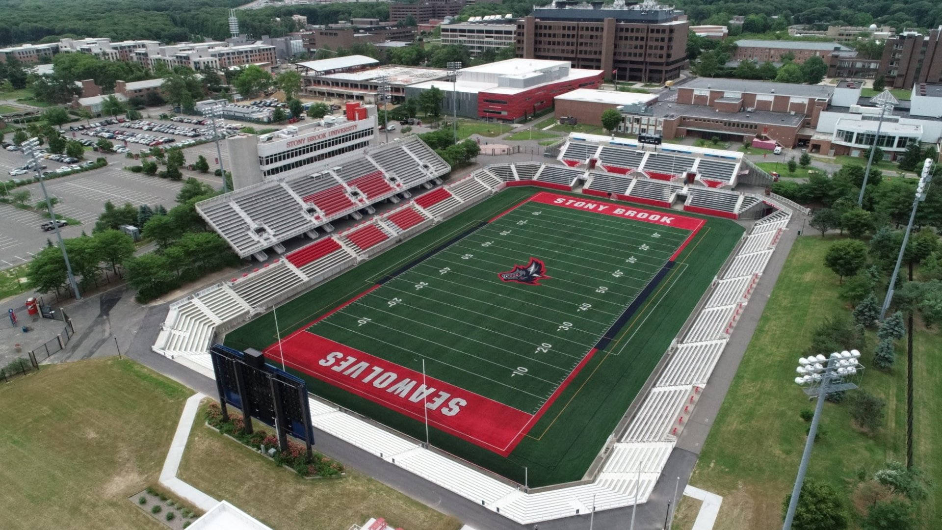 Football Field 桌布 壁紙 — 石溪大學足球場的鳥瞰圖，綠色球場、紅色端區和空蕩蕩的看台，周圍環繞著校園建築和樹木。