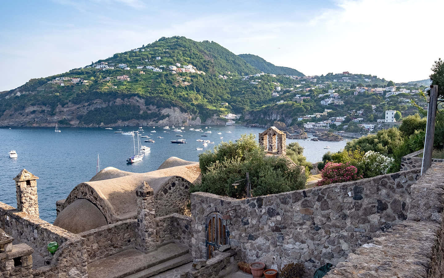 Italian Island 桌布 壁紙 — 石牆露台俯瞰陽光普照的海灣，海灣上有船隻和梯田山坡村莊，天空清澈湛藍。
