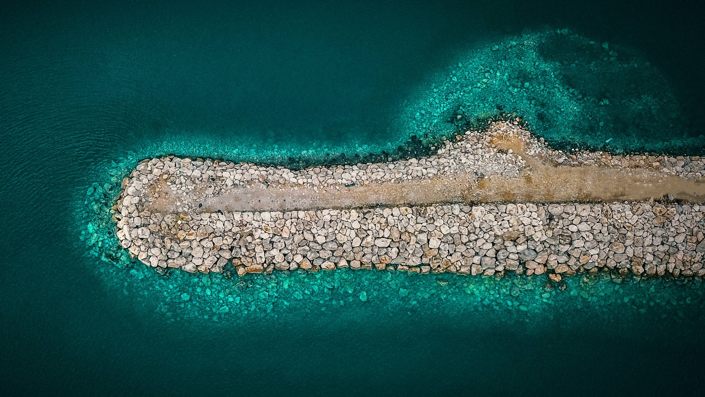 海洋藍 桌布 壁紙 — 石造防波堤延伸至充滿活力的綠松石色海水中的航拍視圖，邊緣清澈淺顯。
