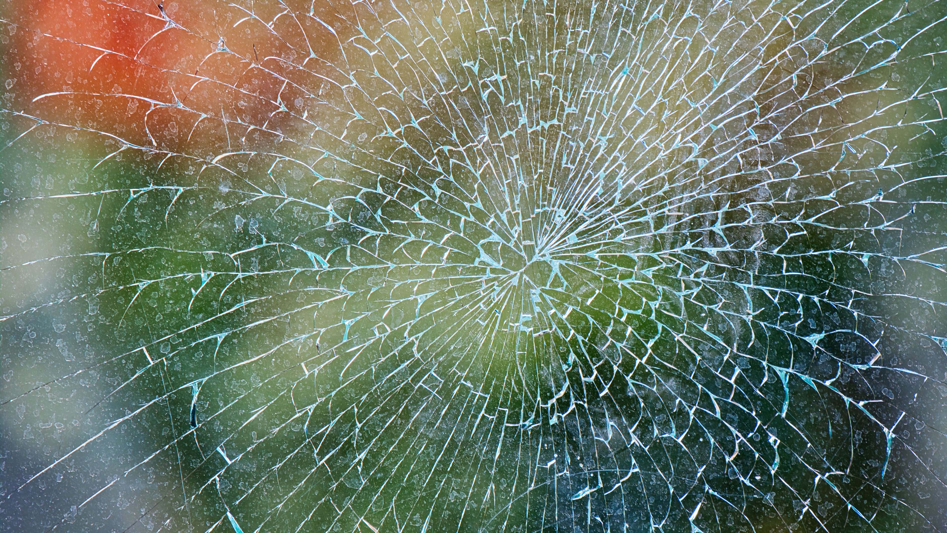 Fracture Glass 桌布 壁紙 — 碎裂玻璃的特寫，帶有放射狀裂紋圖案，顯露出模糊的綠色和橙色背景。