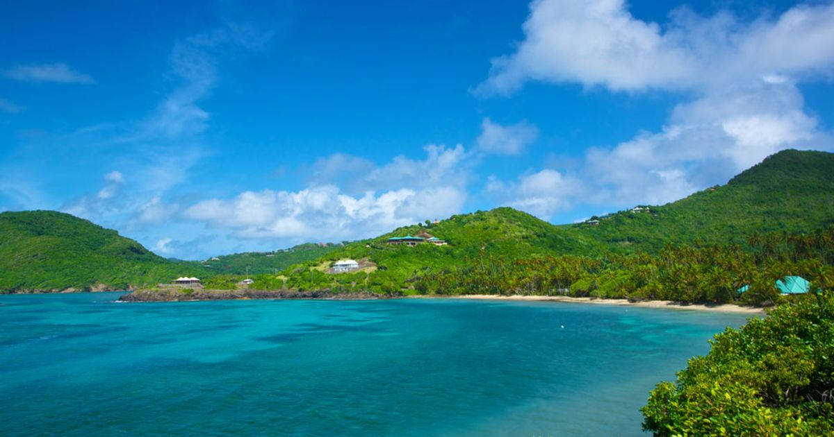 St Vincent And The Grenadines 桌布 壁紙 — 碧綠色的海水拍打著沙灘，沙灘被鬱鬱蔥蔥的綠色山丘環繞，明亮的藍天下飄著蓬鬆的白雲。