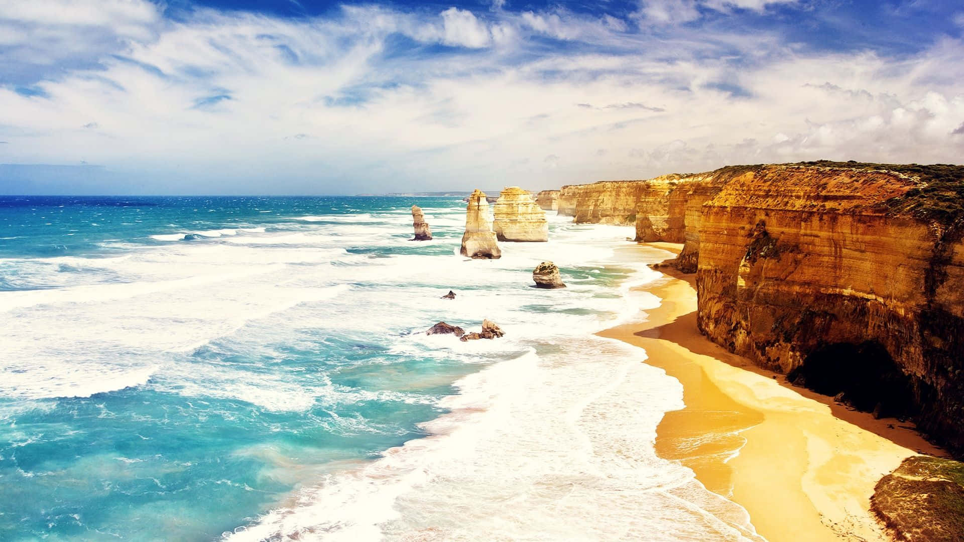 Australia Beach 桌布 壁紙 — 碧綠色的海浪拍打著金色的沙灘，旁邊是高聳的石灰岩懸崖，上方是晴朗的藍天，點綴著幾朵白雲。
