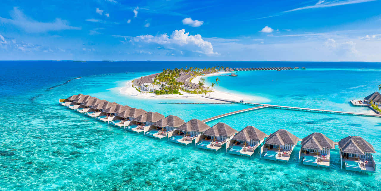 Maldives Island 桌布 壁紙 — 碧藍天空下，綠松石色潟湖上建有水上平房和白色沙島的鳥瞰圖。