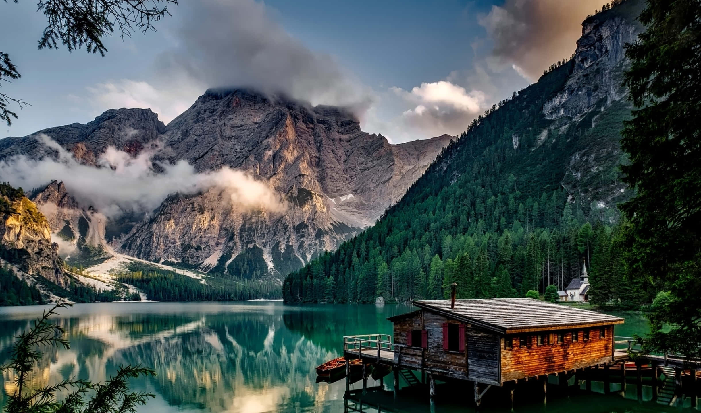 Italy Aesthetic 桌布 壁紙 — 碼頭上的木屋倒映在碧綠色的水中，周圍環繞著高聳雲霧繚繞的山脈和茂密的常綠森林。