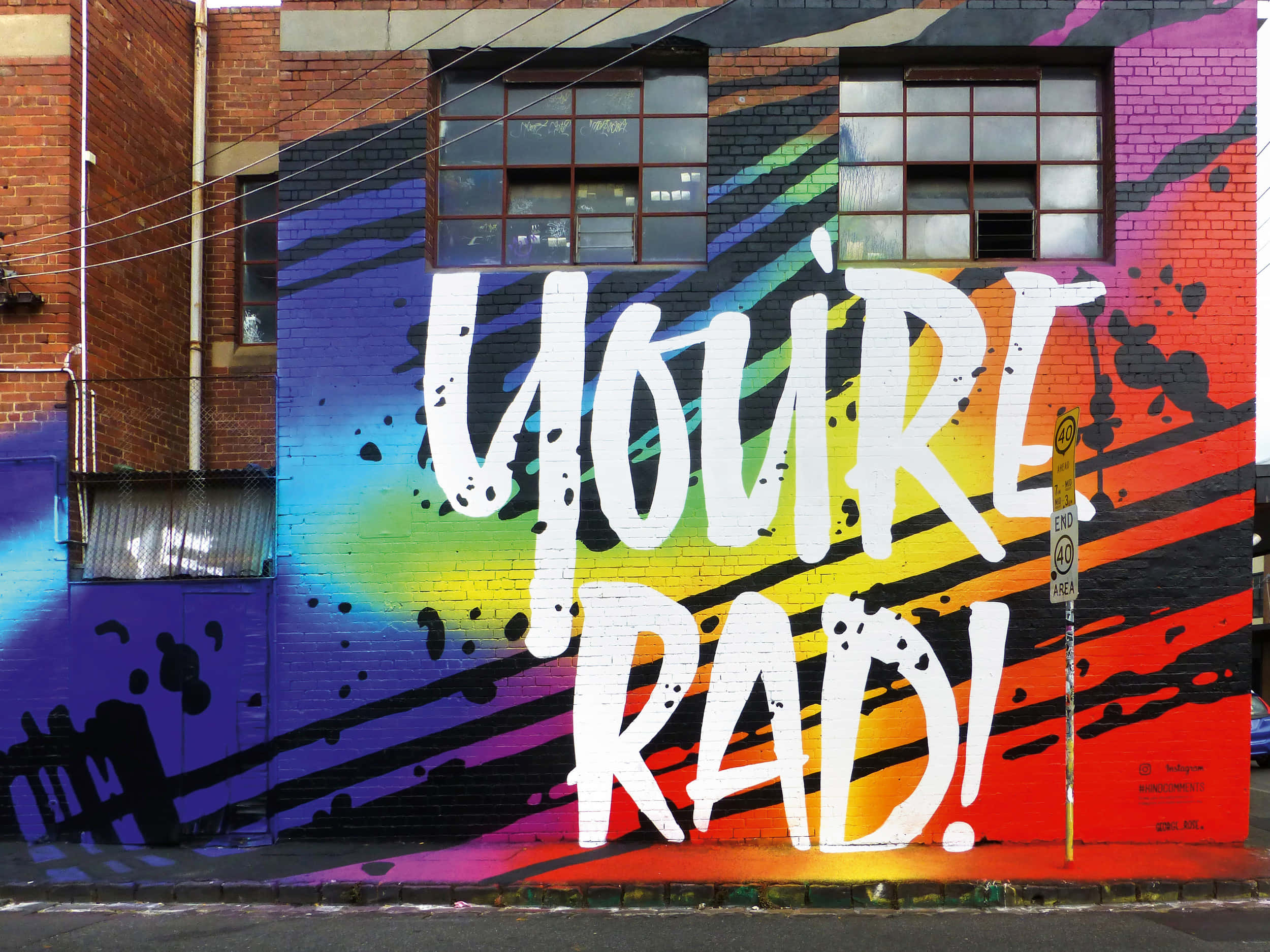 Graffiti Art 桌布 壁紙 — 磚牆上充滿活力的塗鴉壁畫，以粗體白色字母寫著「YOU'RE RAD!」，背景是彩虹條紋和黑色潑濺。