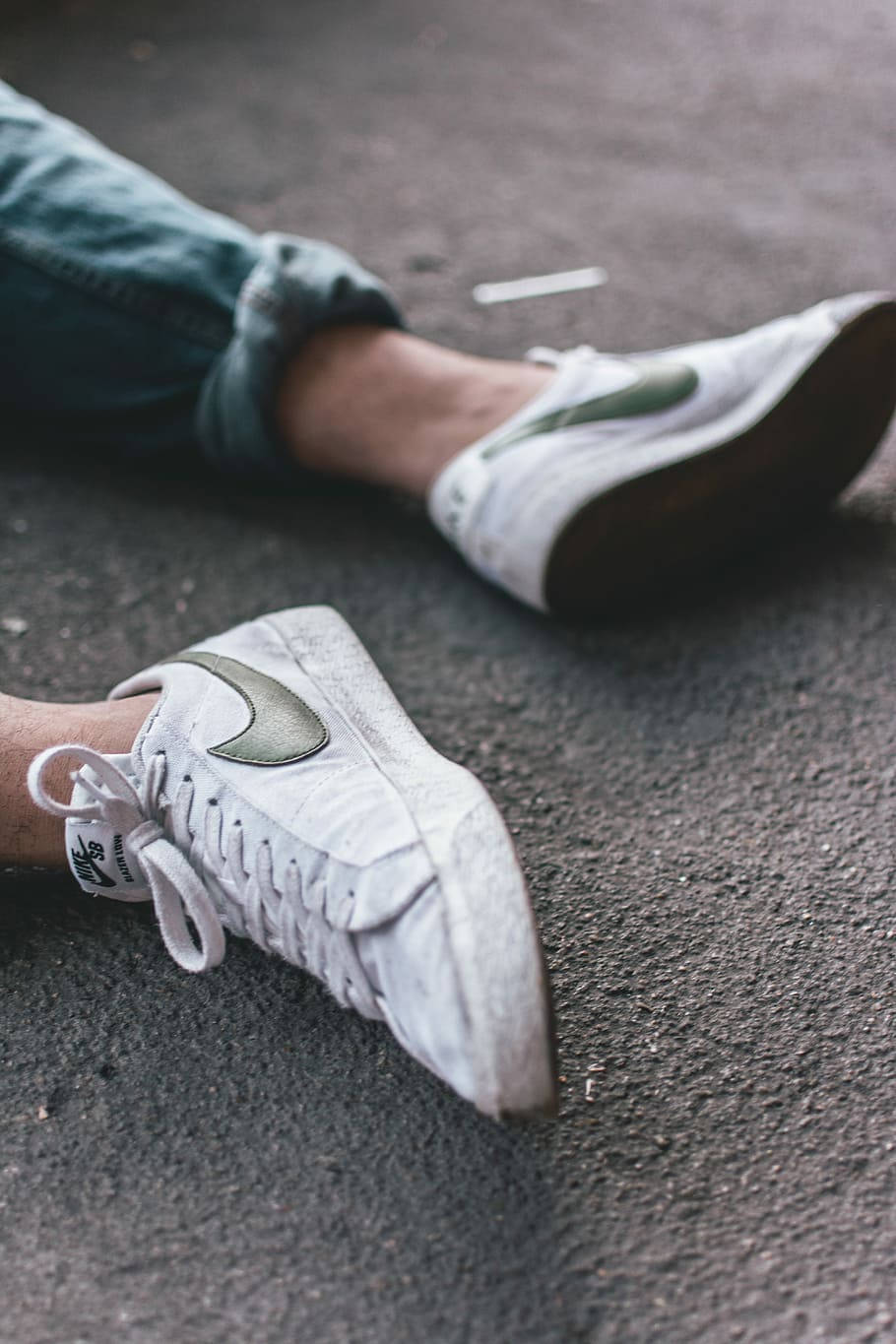 Nike Shoes 桌布 壁紙 — 磨損的白色運動鞋帶有綠色勾勾，放在粗糙的瀝青上，穿著捲起牛仔褲的雙腿放鬆地躺著。