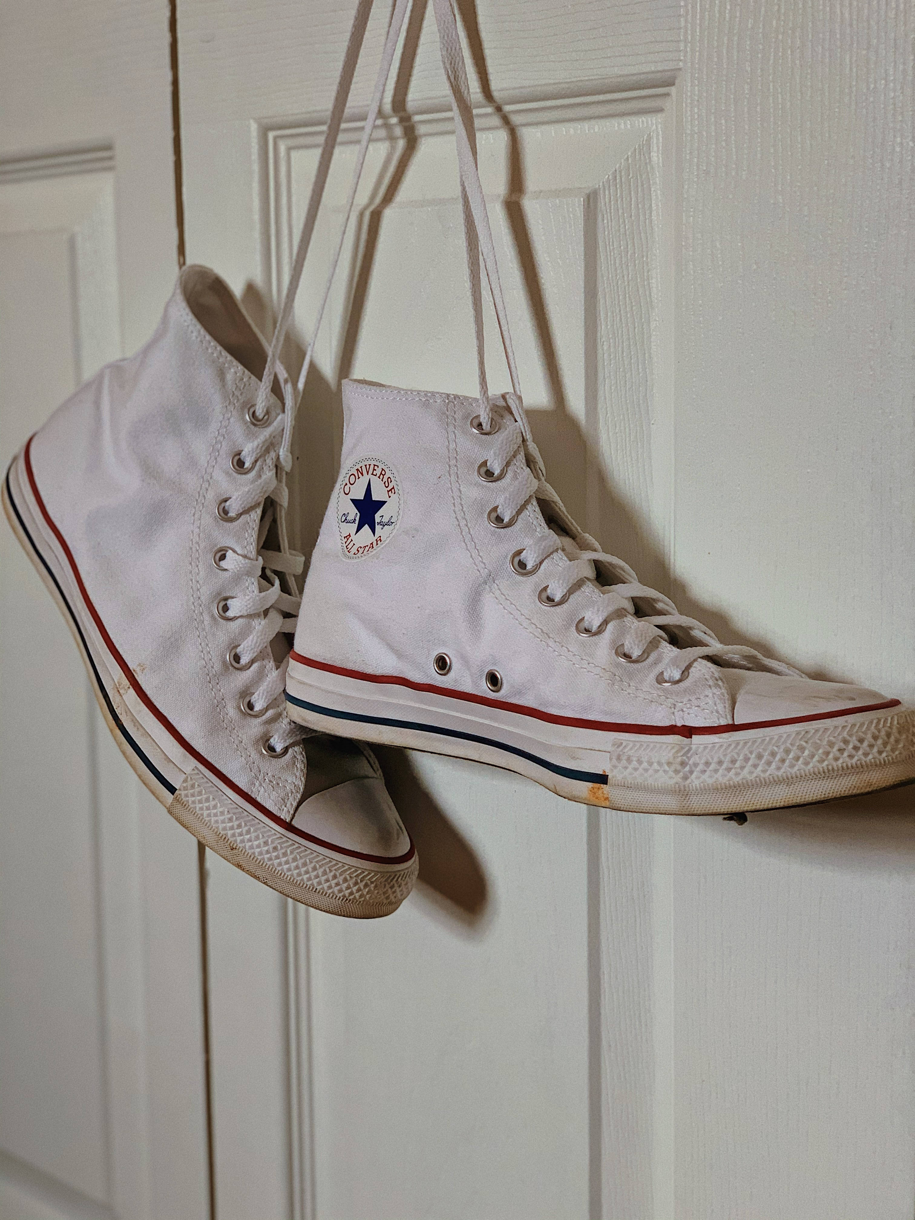 Converse 桌布 壁紙 — 磨損的白色高筒Converse運動鞋用鞋帶掛在白色鑲板門上，顯示磨損的鞋底和經典的紅藍飾邊。