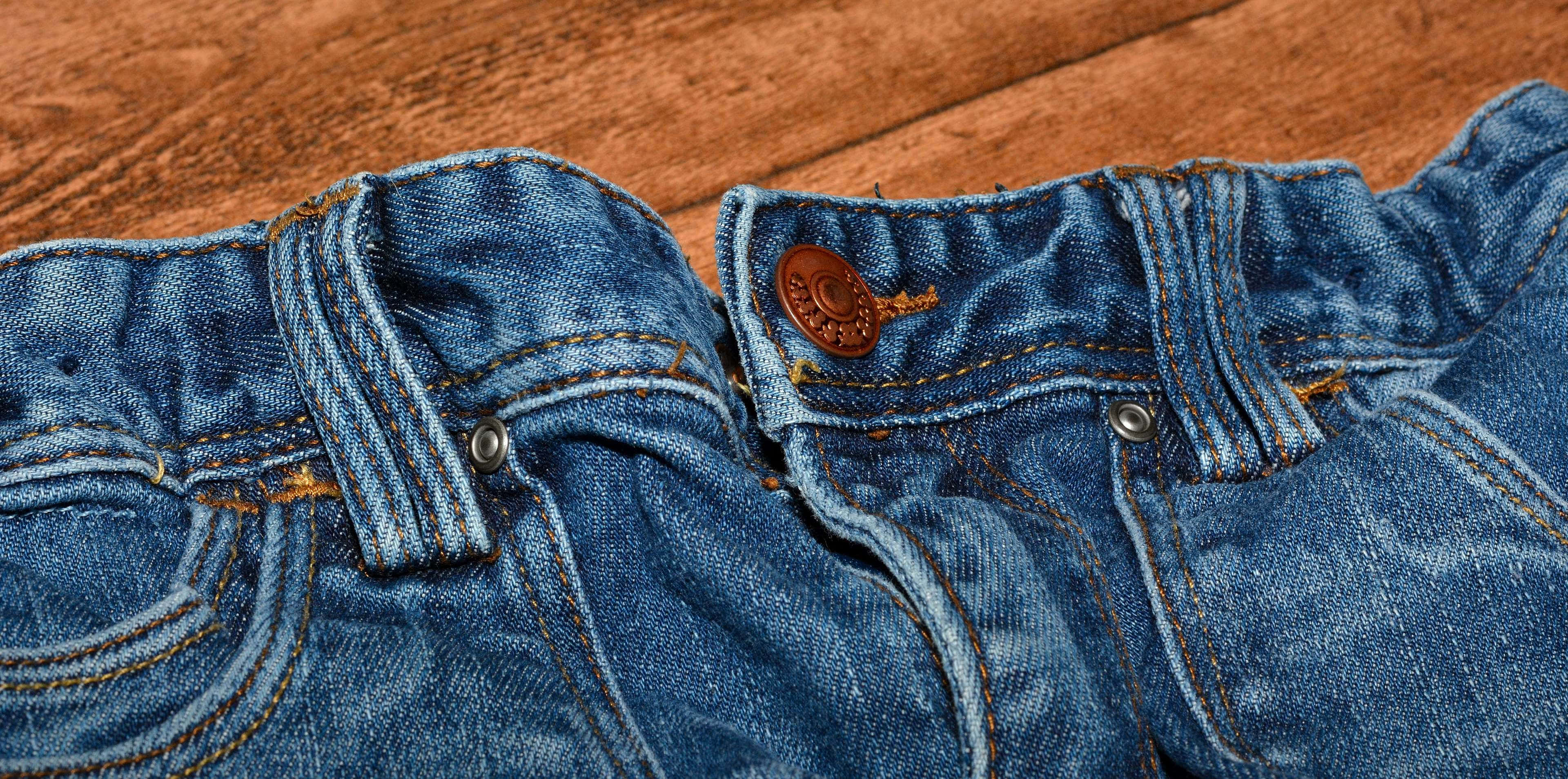 Denim 桌布 壁紙 — 磨損的藍色牛仔褲特寫，有棕色縫線和鈕扣，放在溫暖的木質表面上。