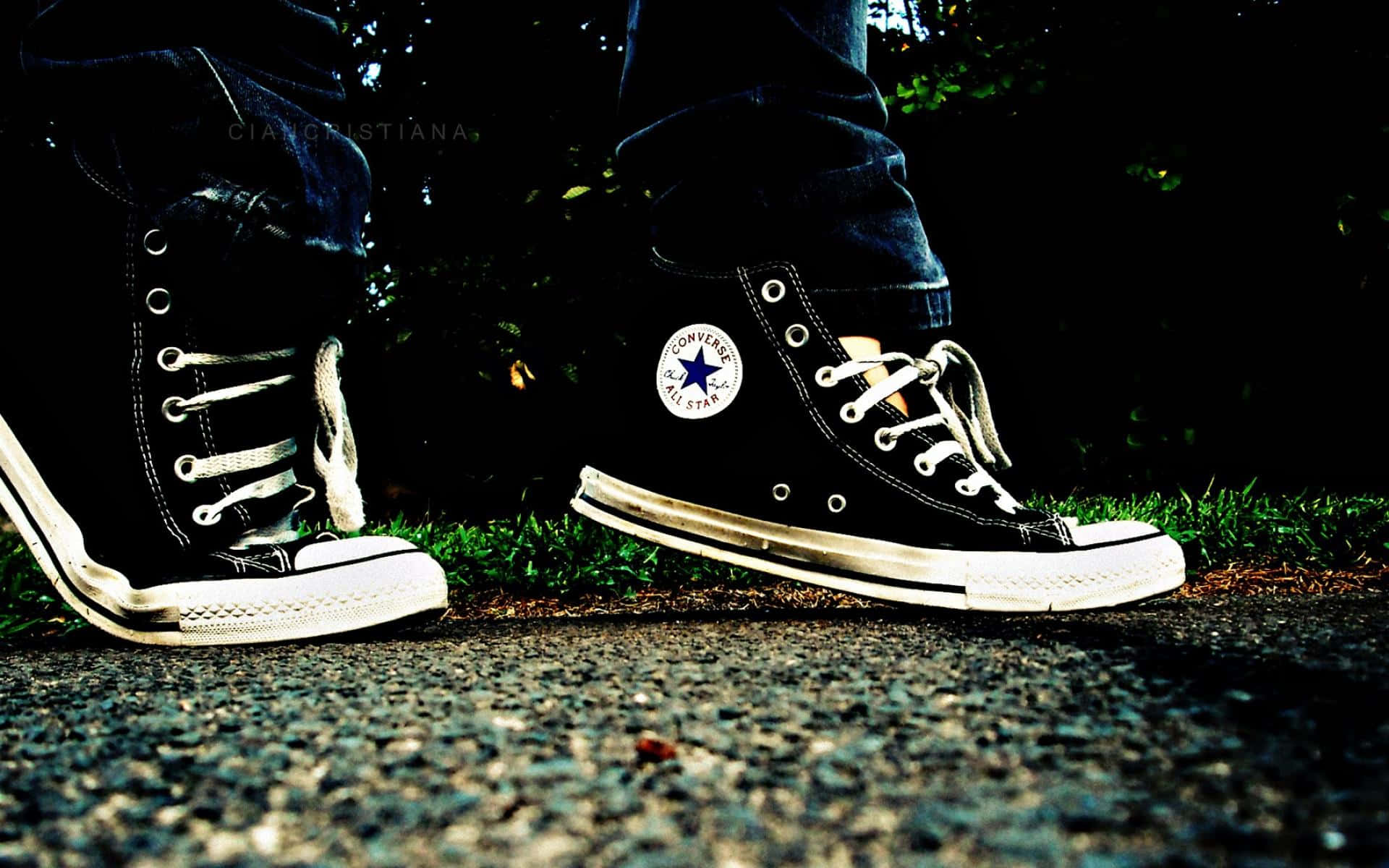 Converse 桌布 壁紙 — 磨損的黑色Converse運動鞋特寫，帶白色鞋帶和鞋底，站在暗色樹葉下的草地附近的礫石上。