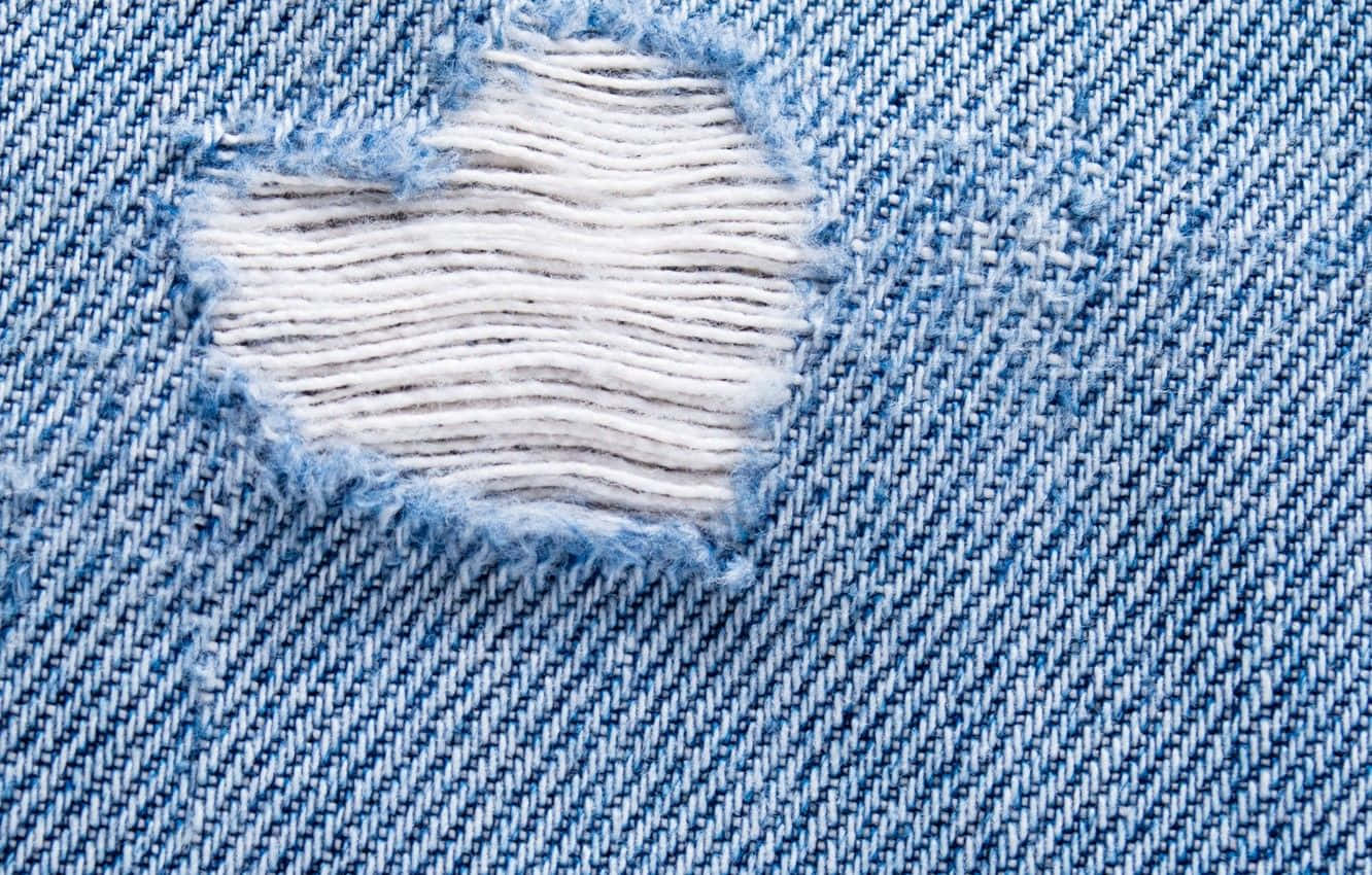 Denim 桌布 壁紙 — 磨損藍色牛仔布料的特寫，有磨損的圓形撕裂，露出下面的白色縫線。